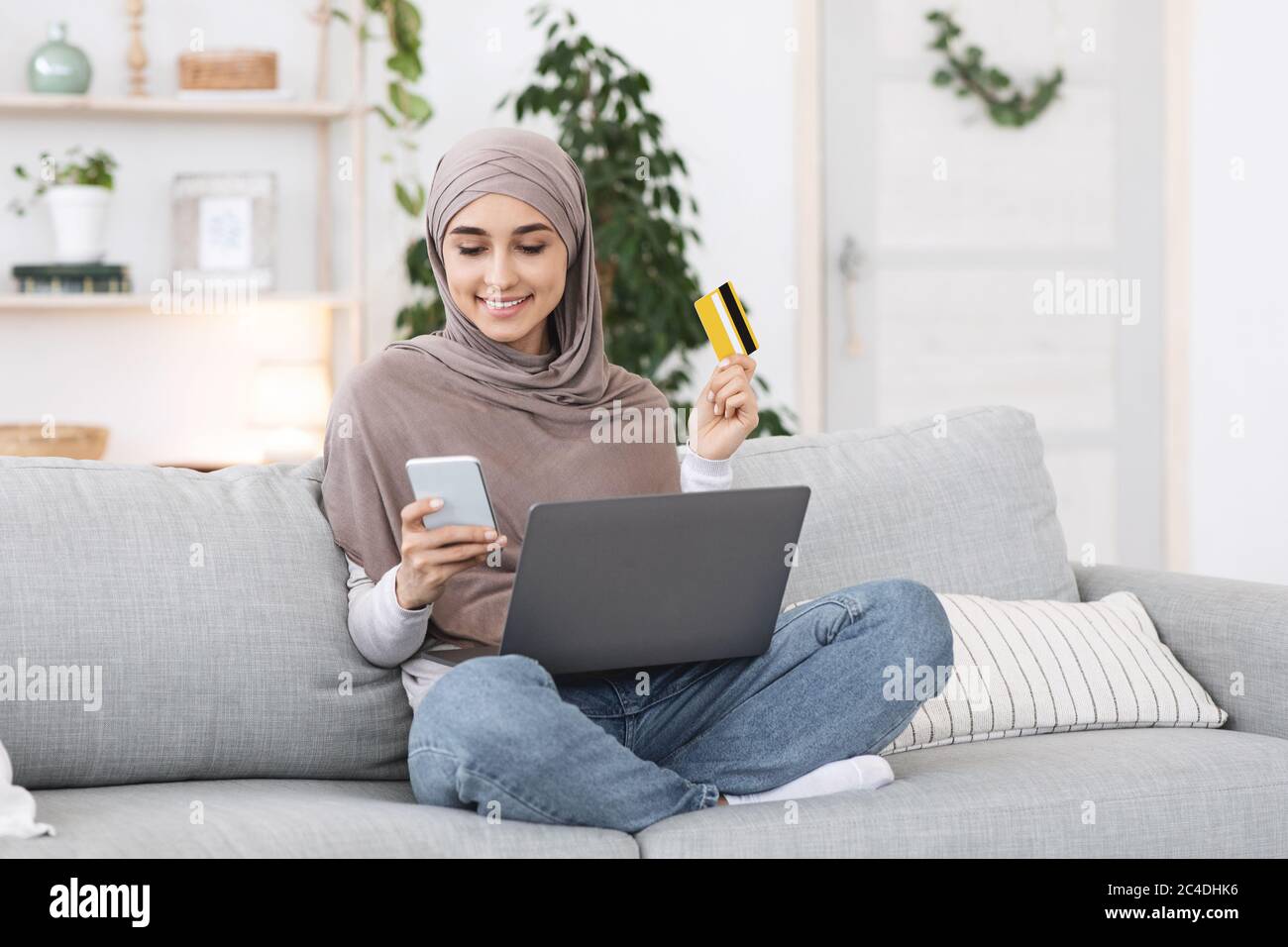 Online Payments. Happy arabic girl using smartphone, laptop and card at home Stock Photo