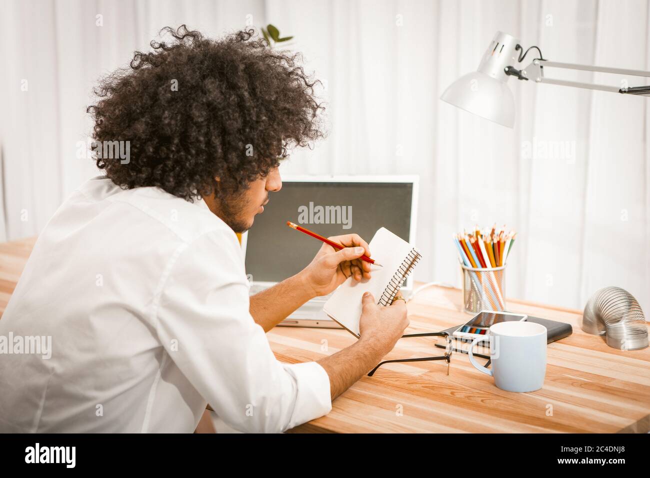 Side view of shaggy man making notes in his paper notebook or scratchpad sitting at wooden table with computer and cup of hot beverage on it. Memory Stock Photo