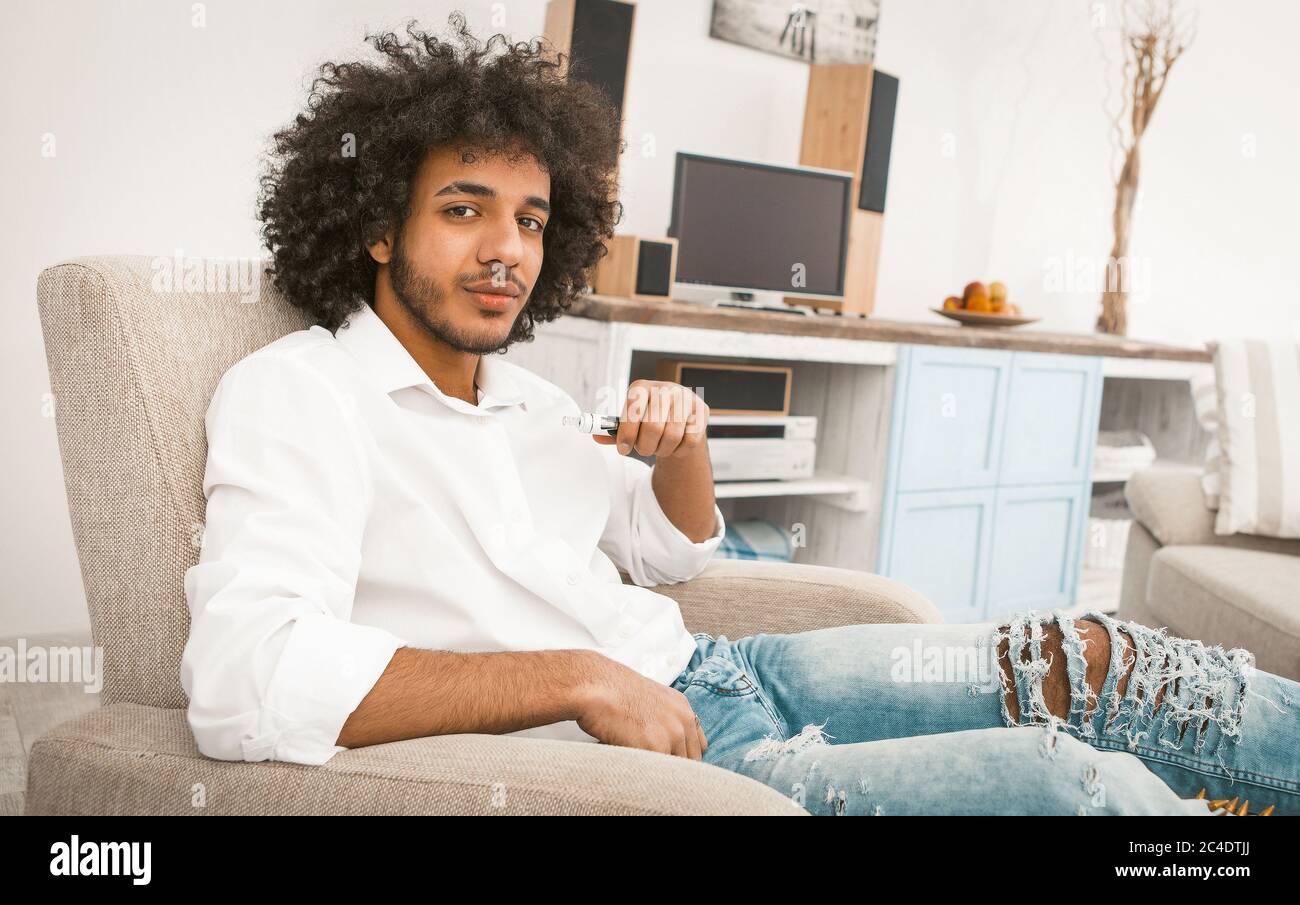 Young handsome guy smoking of electronic or e-cigarette while sitting at armchair on modern home interior background. Tinted image Stock Photo
