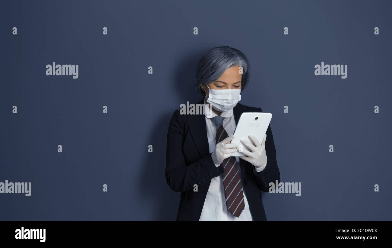 Mature woman works with digital tablet. Gray-haired businesswoman in black suit using electronic gadget standing against on gray office wall Stock Photo