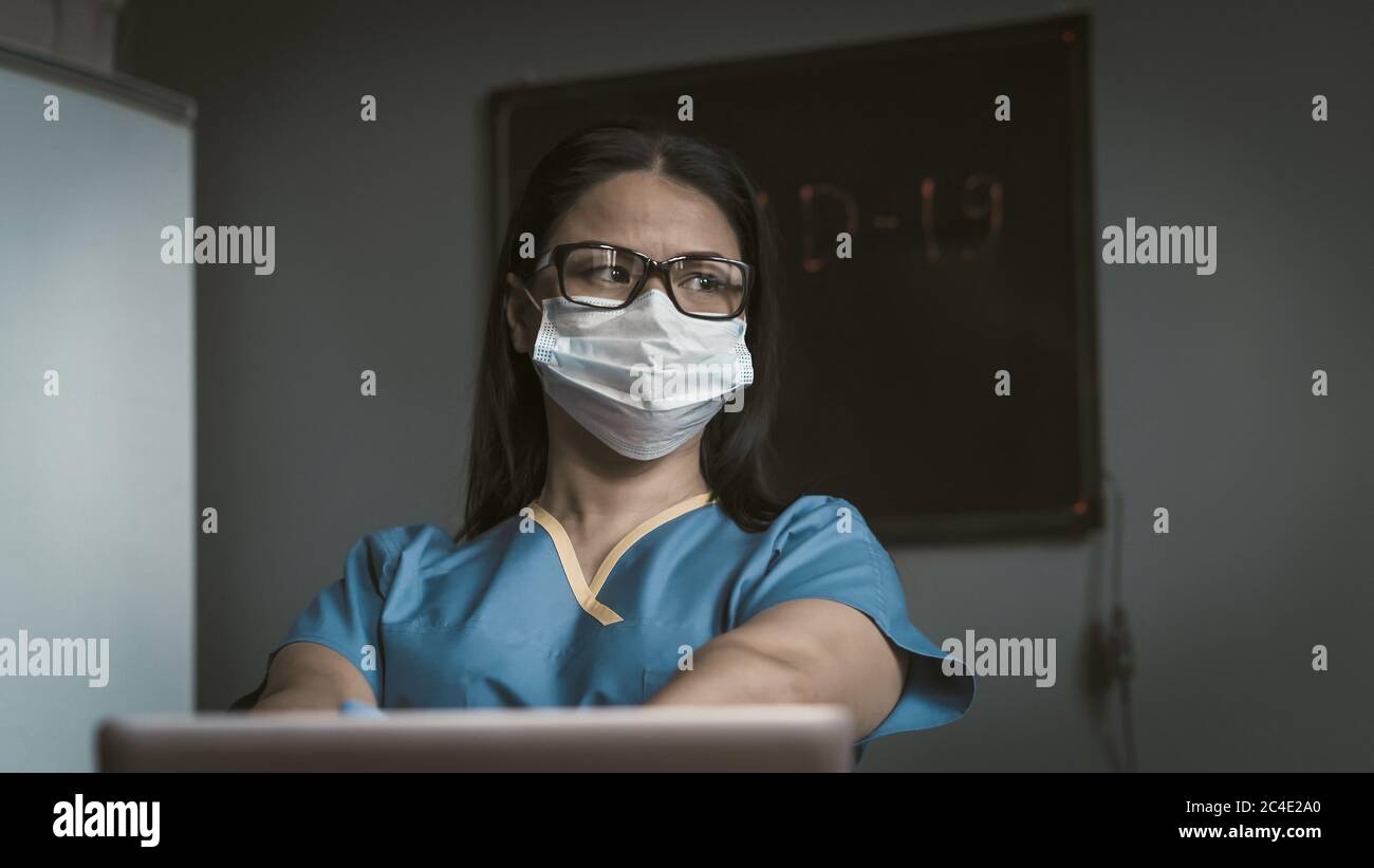 Tired medic looks thoughtfully at side. Pensive female doctor thinks sitting at desk with laptop on it. Pandemic concept. Toned image Stock Photo