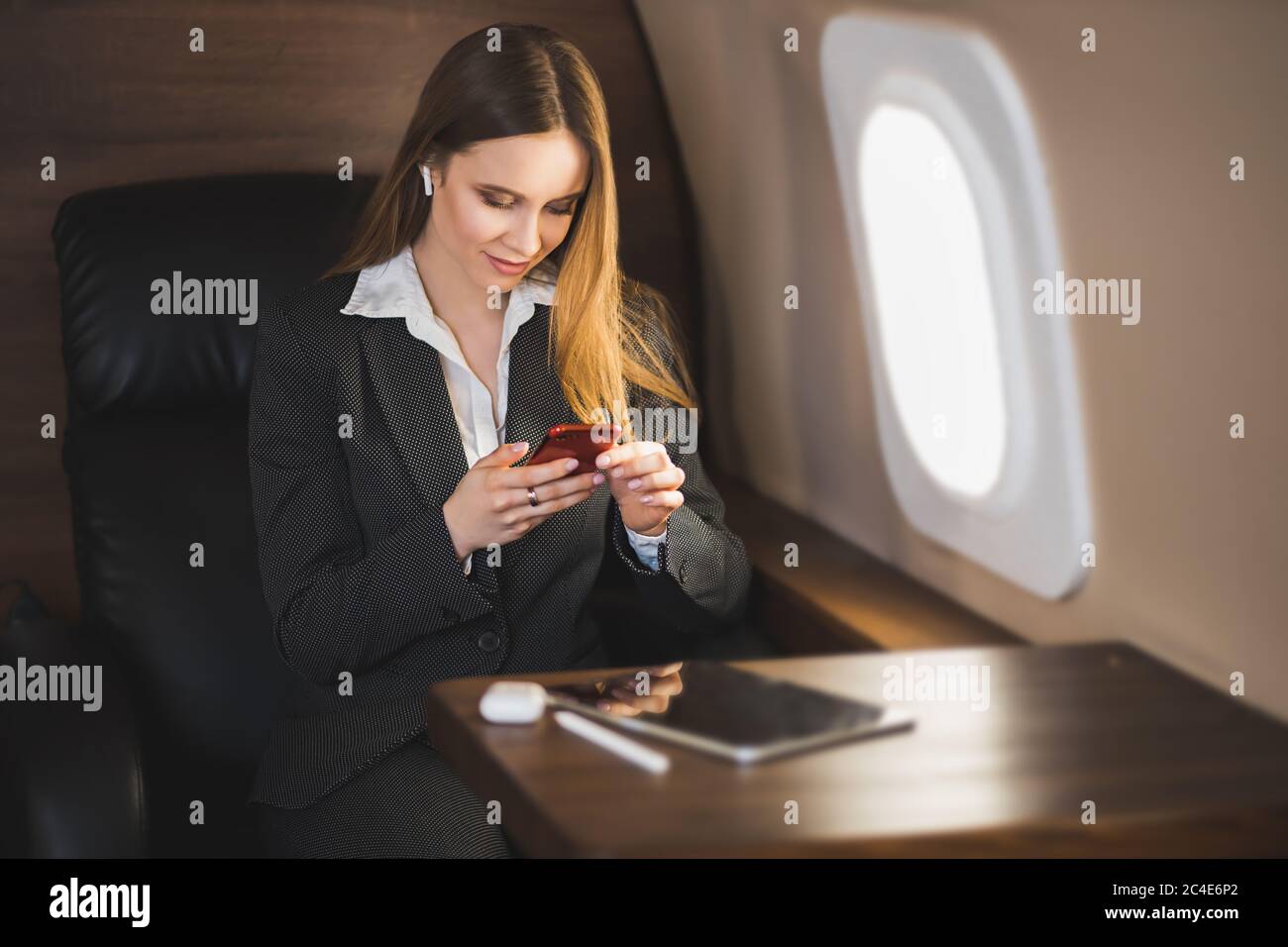 Lovely business lady in white blouse and formal suit, sitting in comfortable seat, looking at screen of smartphone, using bluetooth earpads during fli Stock Photo