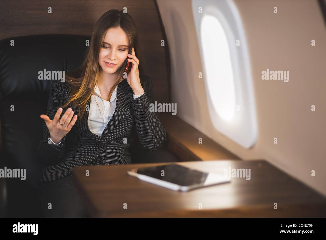 Cheerful happy brown haired business lady in white blouse and formal suit, sitting in comfortable seat, laughing talking on mobile phone during flight Stock Photo