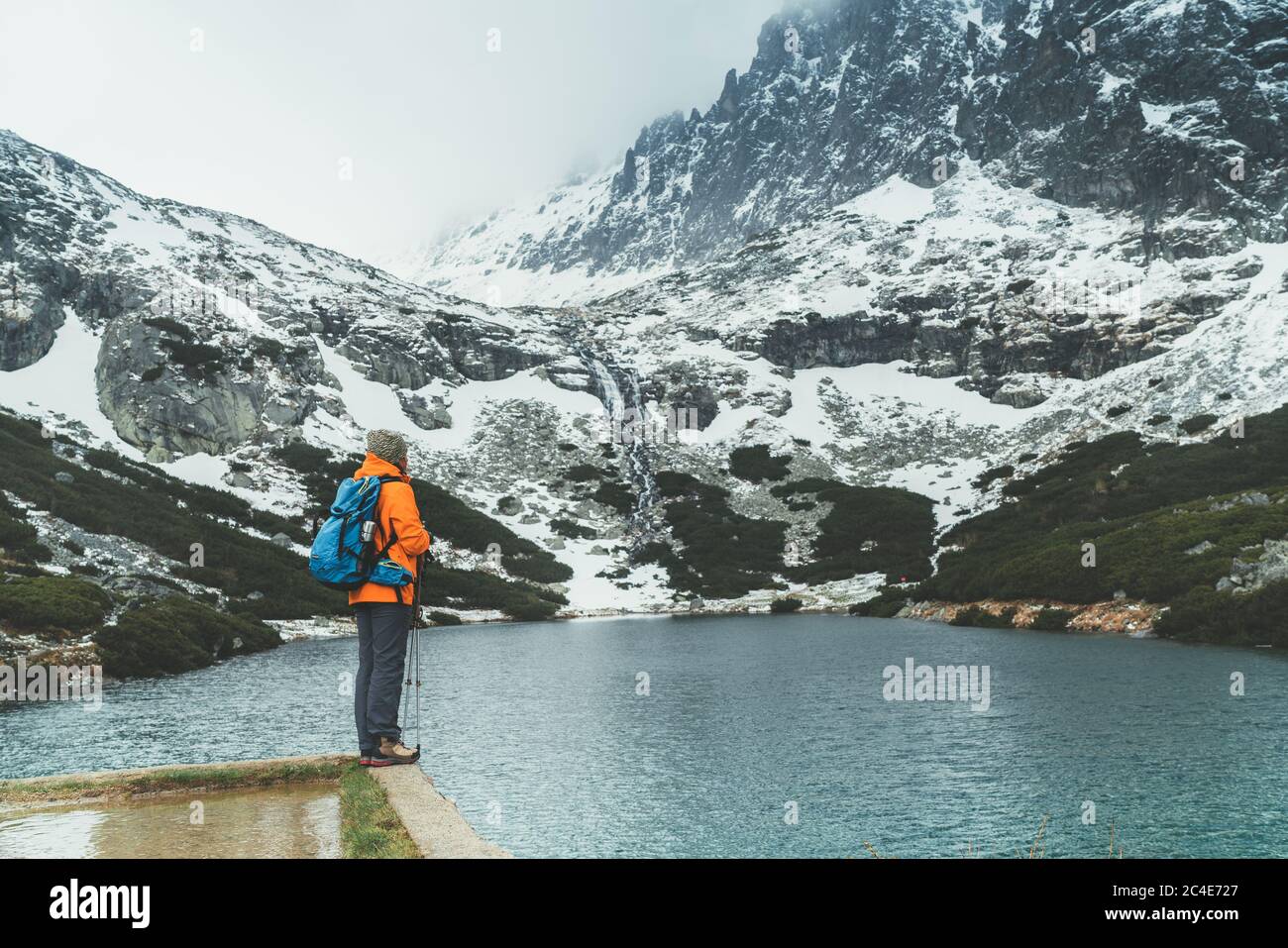 Dressed bright orange jacket female backpacker enjoying the Velicke pleso (mountain lake) view as she have mountain walk in Velicka valley in High Tat Stock Photo