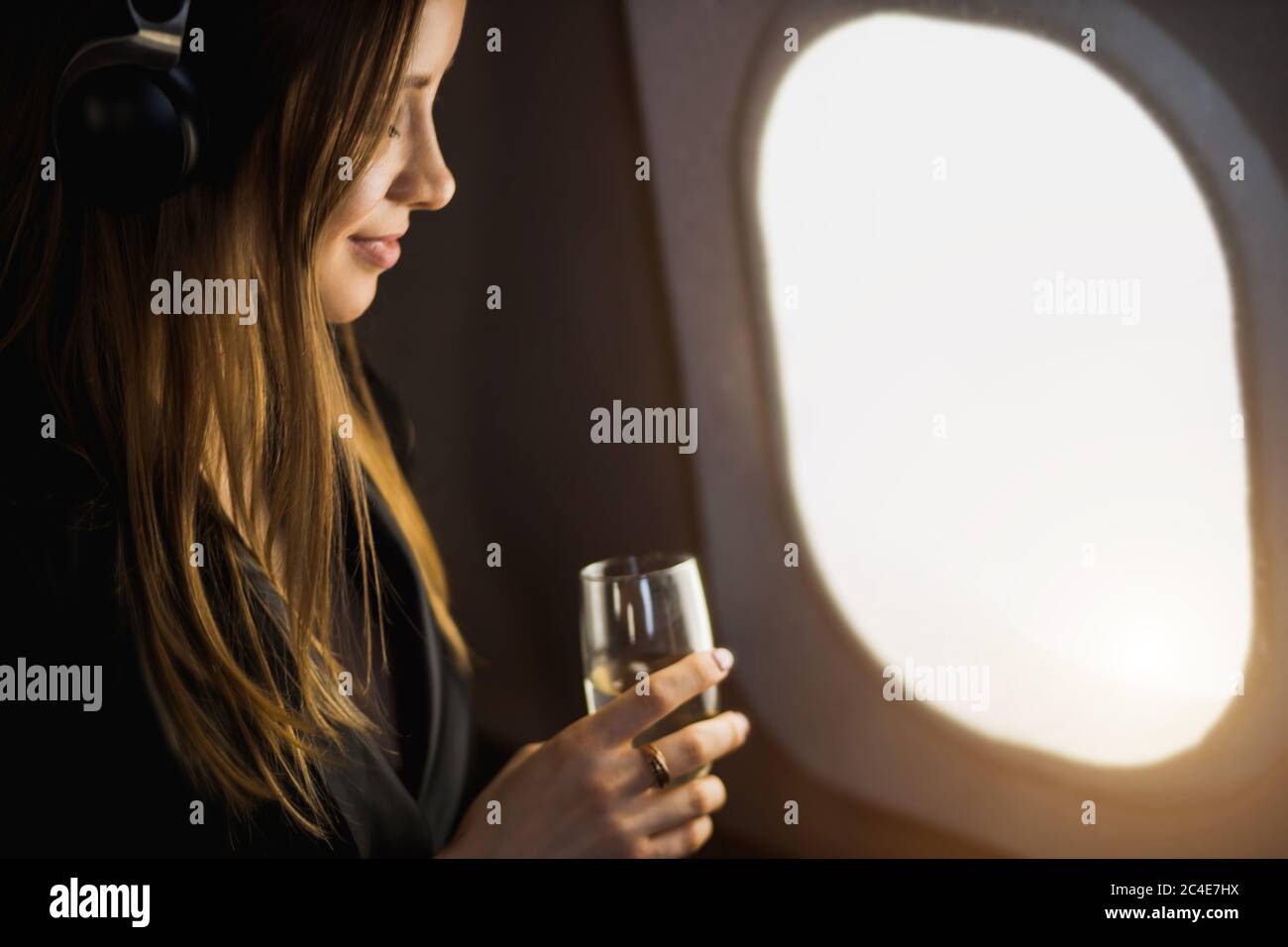 closeup shot of happy caucasian girl with long blonde hair, in black jacket dress, listening to audiobook with headphones, holding glass of champagne Stock Photo