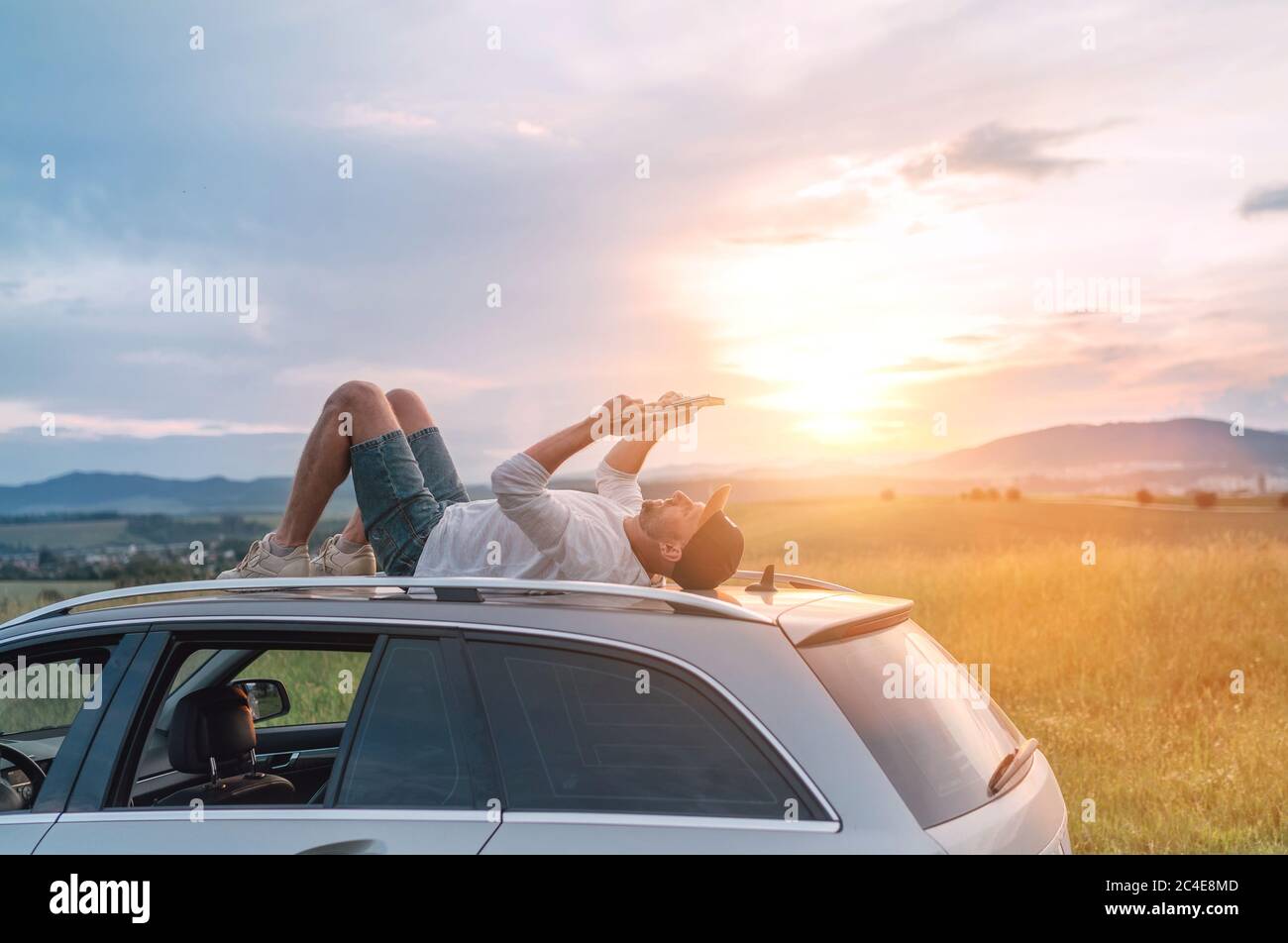 Mature age Man lying on car roof and reading the paper bestseller book. He stopped his auto on high grass meadow with beautiful valley view before the Stock Photo