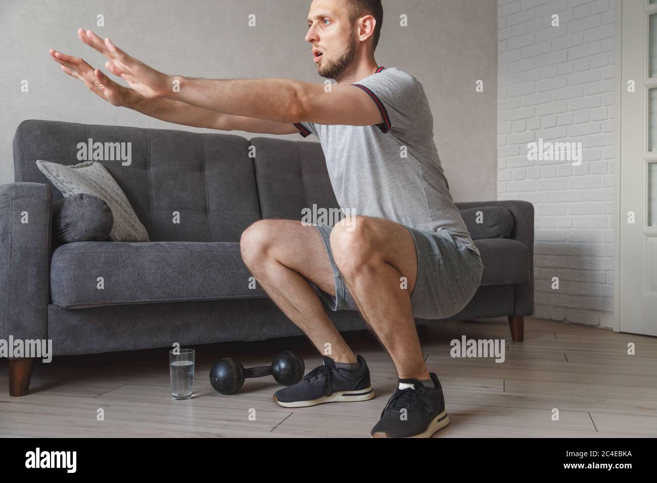Young ordinary man go in for sport at home. Real picture of regular guy doing squats with stretch hands forward. Beginner or amateur has workout in Stock Photo
