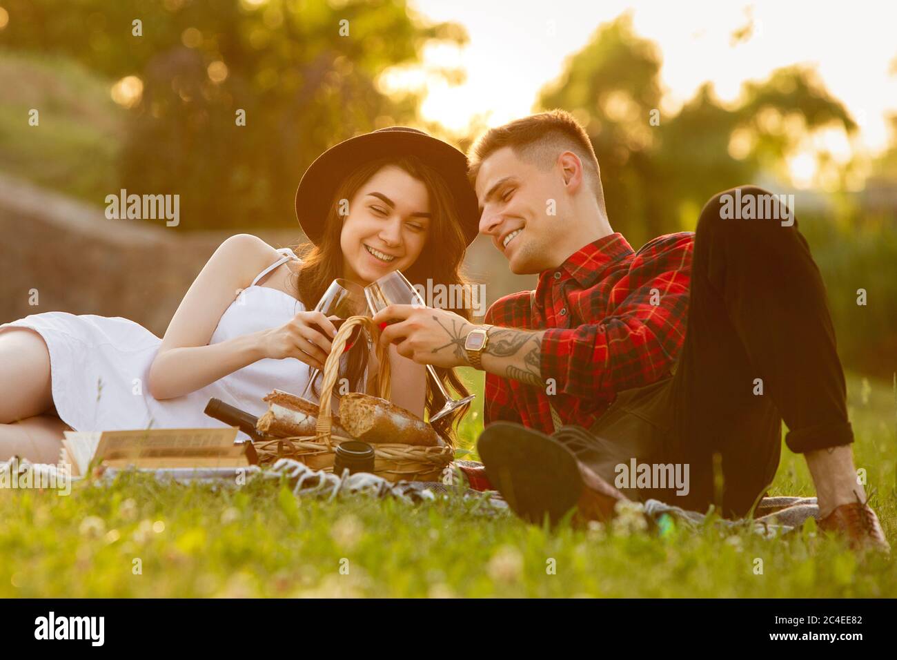 Clinking glasses with wine. Caucasian young couple enjoying weekend in the park on summer day. Look lovely, happy, cheerful. Concept of love, relationship, wellness, lifestyle. Sincere emotions. Stock Photo