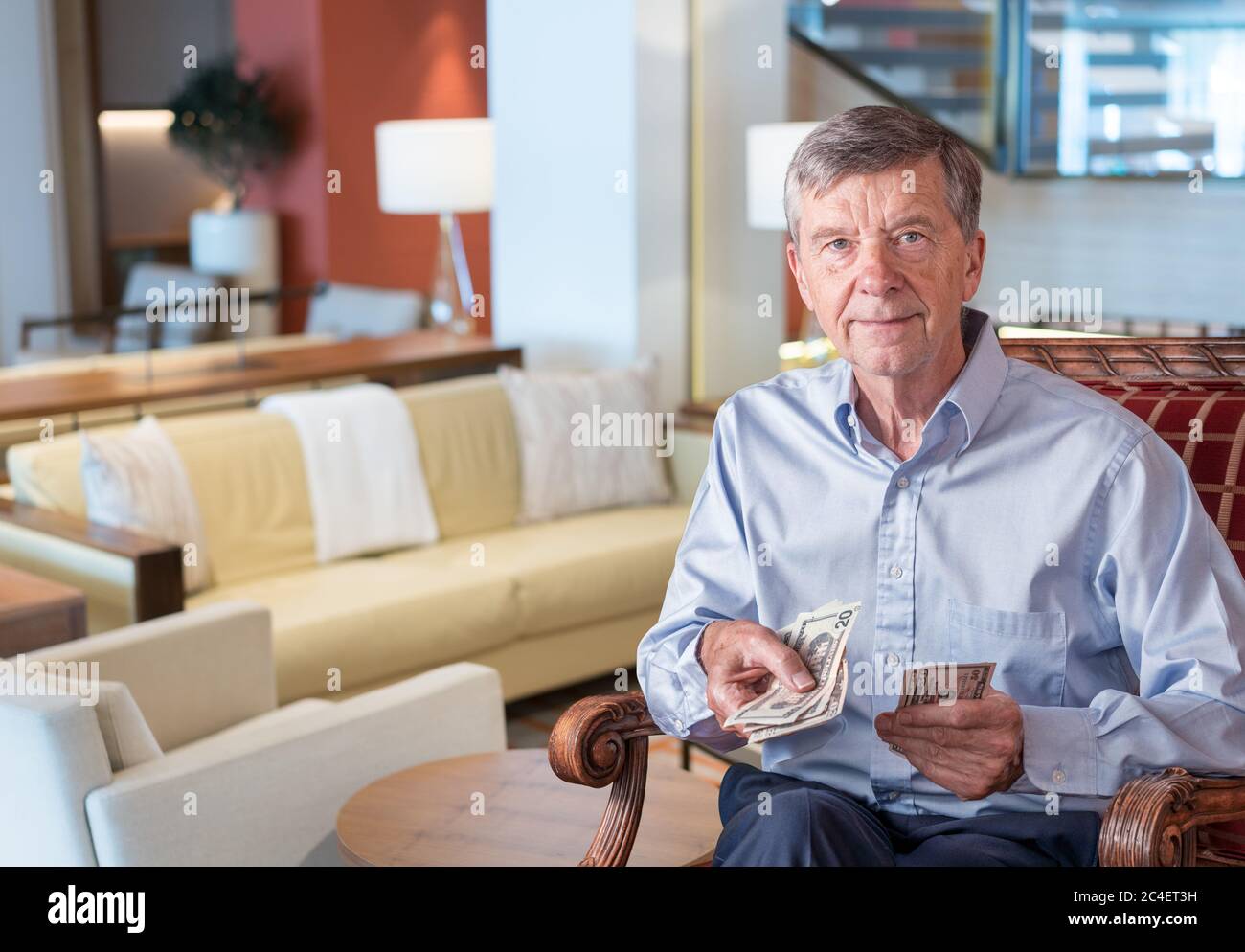 Senior caucasian man holding some US dollar bills as if handing them to the viewer. Composite into upmarket lounge of hotel or cruise ship Stock Photo