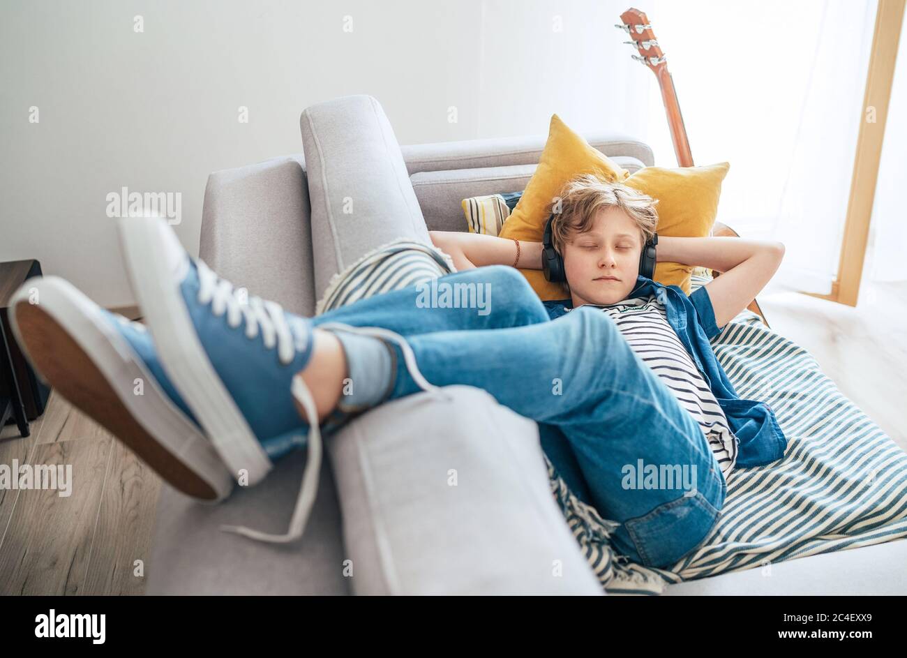 Sleeping preteen boy lying at the home living room filled with sunlight on the cozy sofa dressed casual jeans and sneakers listening to music Stock Photo