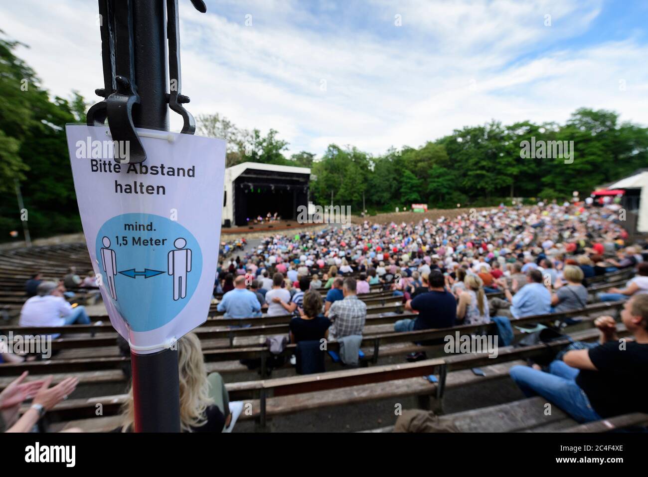 26 June 2020, Saxony, Dresden: A sign with the inscription 'Please keep your distance' hangs on a lamp post while the audience watches the performance of the cabaret group 'Zwinger Trio' with the actors Tom Pauls (l-r), Peter Kube and Jürgen Haase on stage in the open-air theatre Junge Garde. A team of private theatres, promoters and artists have joined forces and from today on, under the title 'By far the best open air theatre in the world', will be staging theatre and concerts in classical amphitheatre architecture. The performances are limited to a maximum of 990 spectators. Photo: Robert M Stock Photo