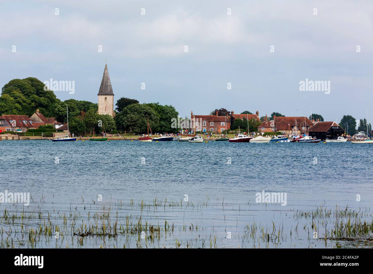 High tide Bosham in West Sussex across Chichester habour on a summers day. Holiday makers and picnickers on the lawn in front of the Holy Trinity. Stock Photo