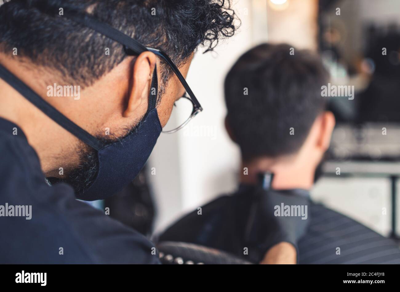 Mulatto barber with a face mask and glasses cut hair on neck and nape of man in barbershop with professional clipper tool. Electric trimmer machine cu Stock Photo