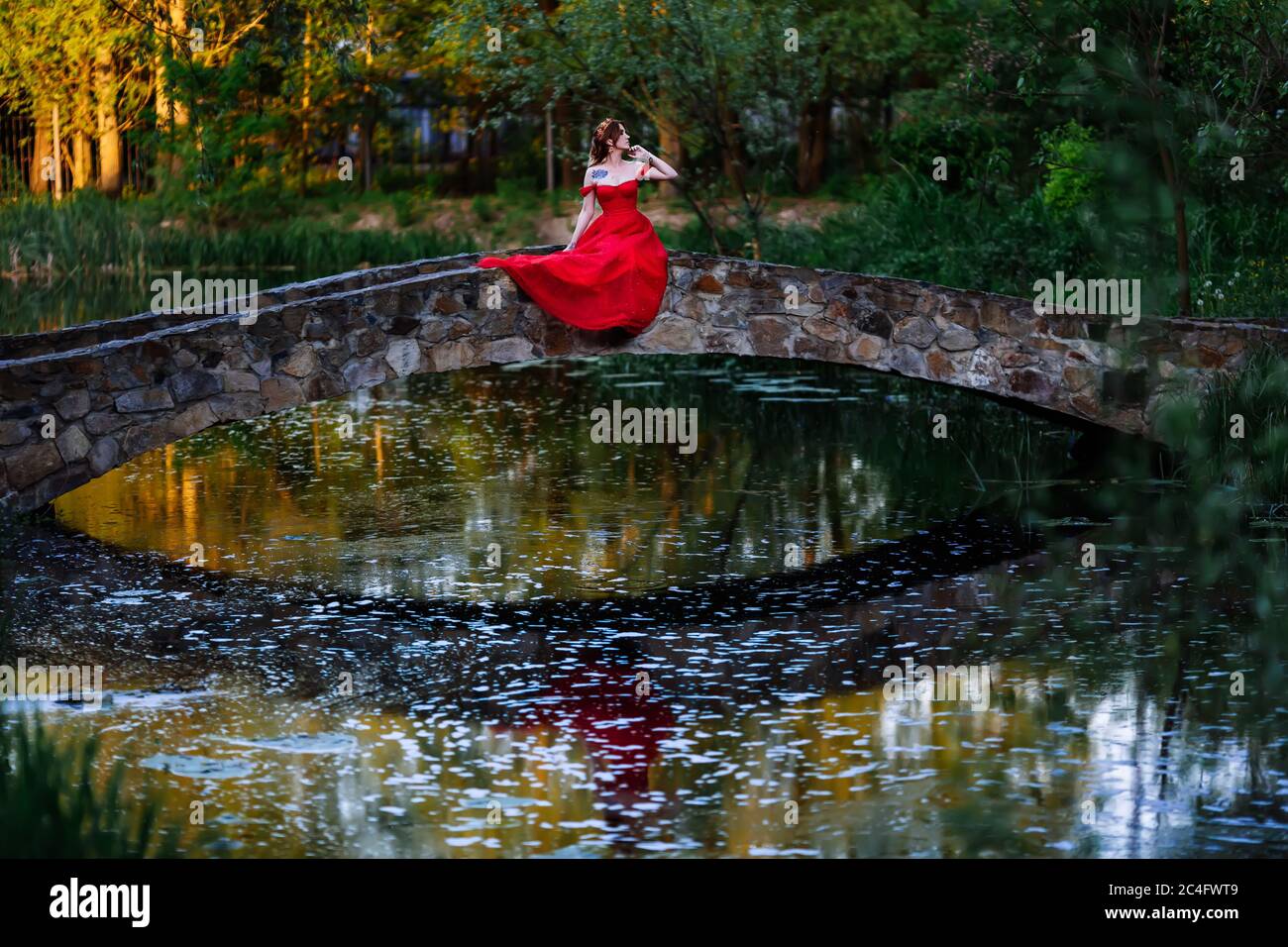 Redhead tattoed woman in red dress sitting on stone bridge Stock Photo
