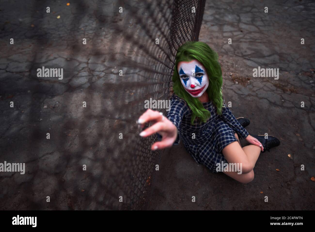 Greenhaired girl in chekered dress with joker makeup sitting near wire mesh fence on the ground. Wide shot. Stock Photo