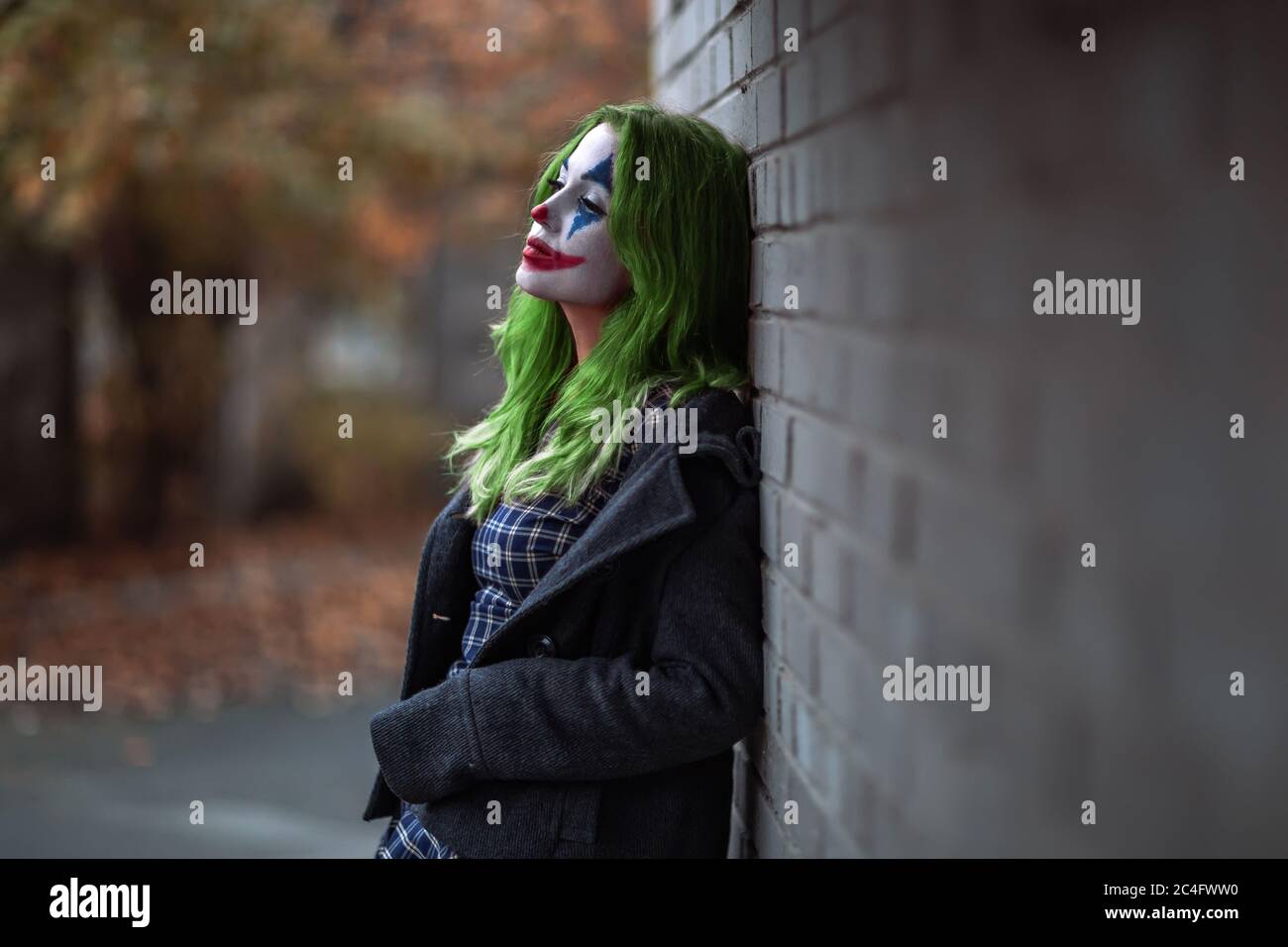 Portrait of a greenhaired girl in chekered dress with joker makeup on a brick wall blurred background. Stock Photo