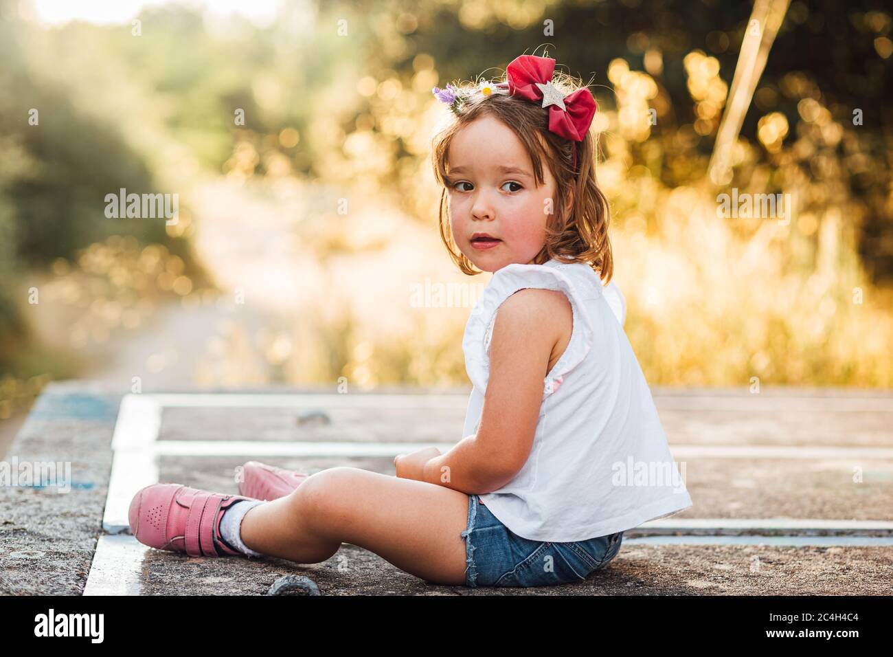 Little blond-haired girl sitting on a concrete platform in the field at sunset Stock Photo