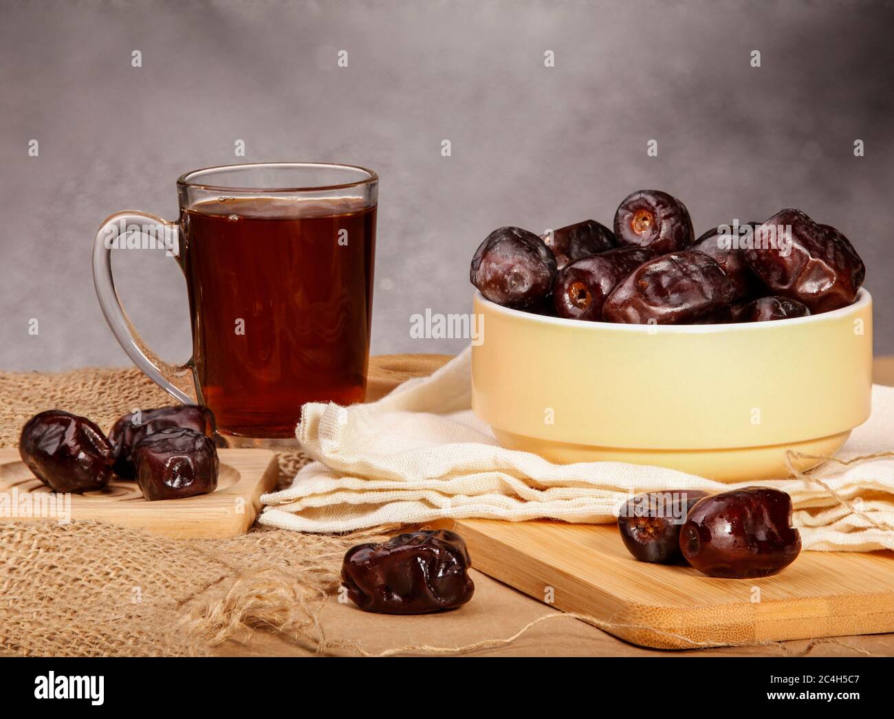 Some dates in a light yellow ceramic bowl and some dates around it on a wooden and sacking surface plus a glass cup of tea. Ramadan dates and tea. Stock Photo