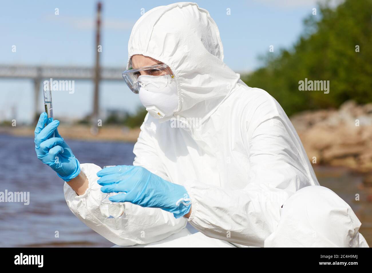 Scientist in protective workwear holding flask with liquid in her hands and examining it outdoors Stock Photo