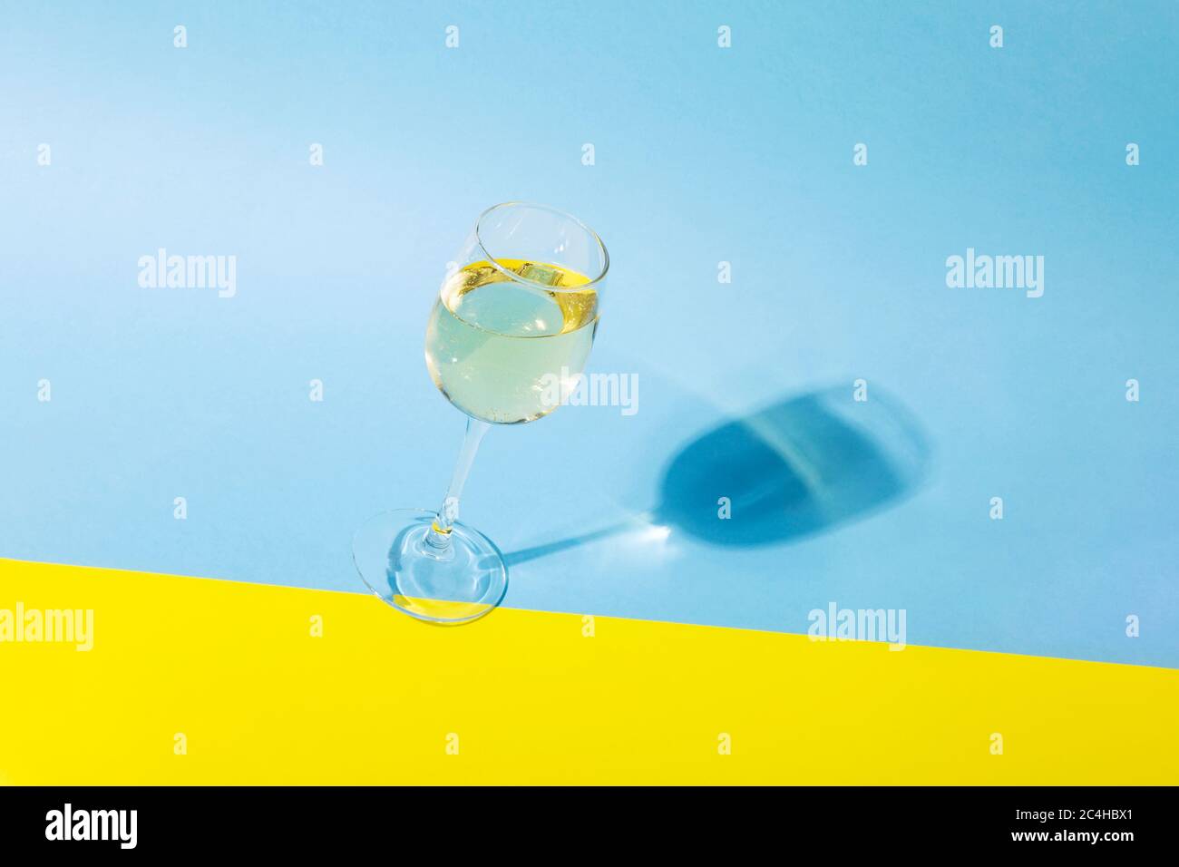 Glass of white wine is tilted on yellow blue background, long shadow from the glass. Shadow effect, hard light Stock Photo