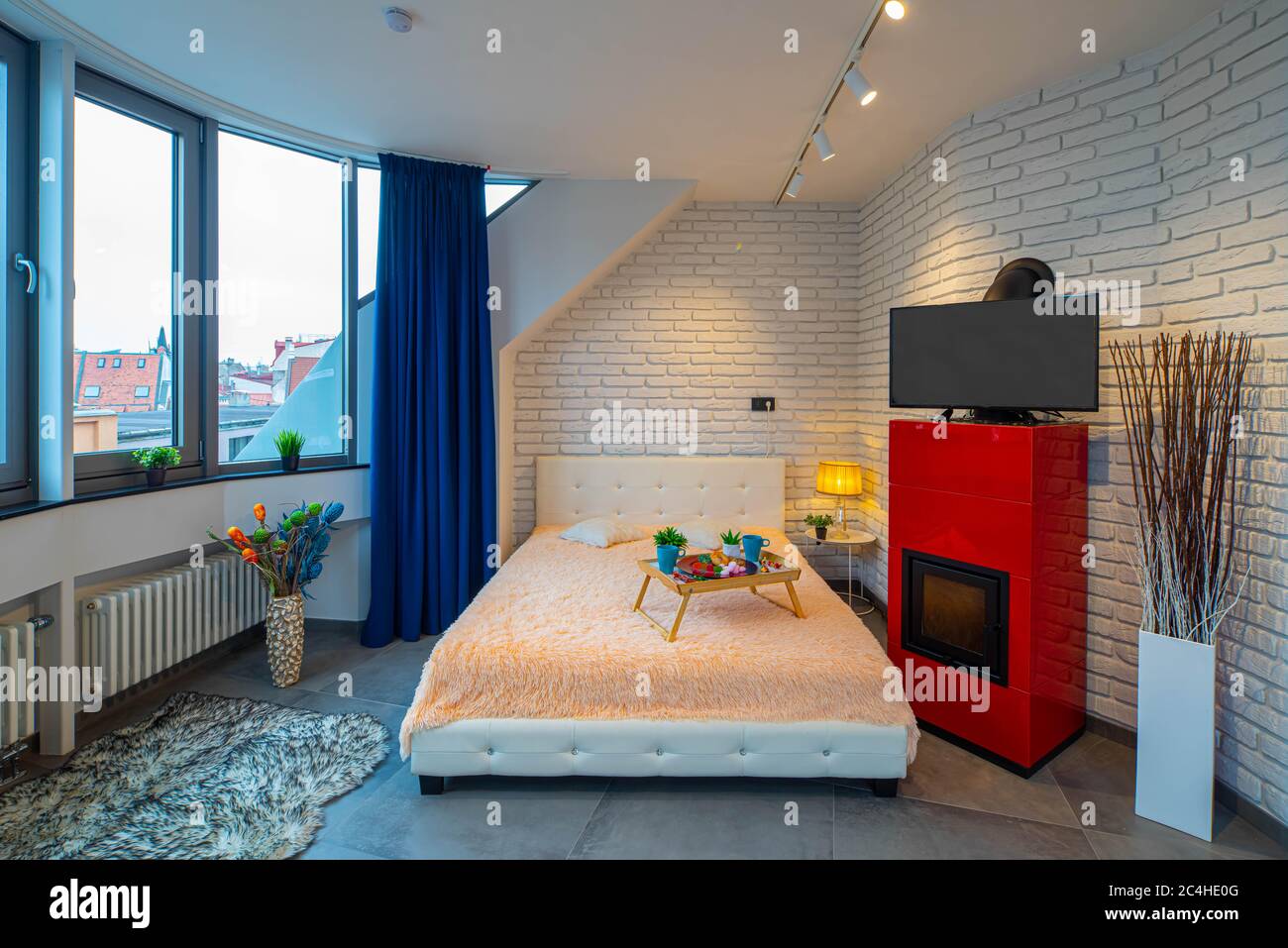 Modern loft interior of luxury studio apartment. Brick wall. Panorama window. Tray with food on the bed. TV on the fireplace. Front view. Stock Photo