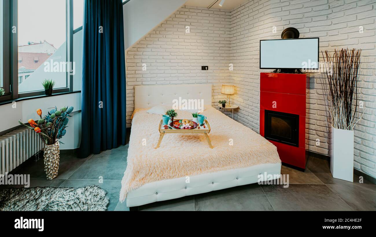 Modern loft interior of luxury studio apartment. Brick wall. Panorama window. Tray with food on the bed. TV on the fireplace. Stock Photo
