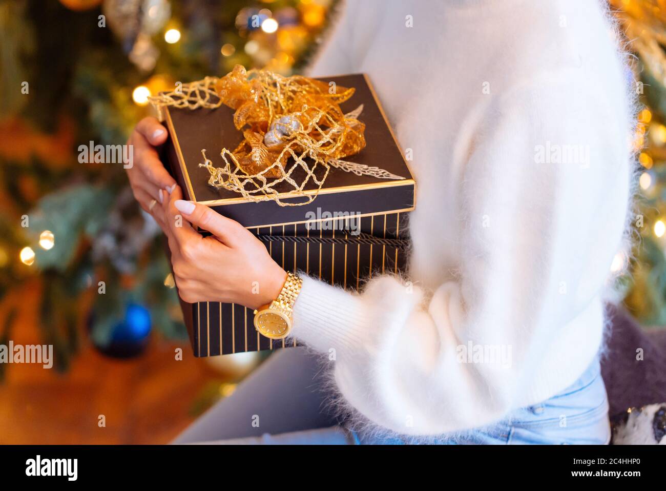 Closeup of female hands holding a present (gift box) in square box, soft focus, Christmas tree on blurred background. New Year, Birthday concept Stock Photo