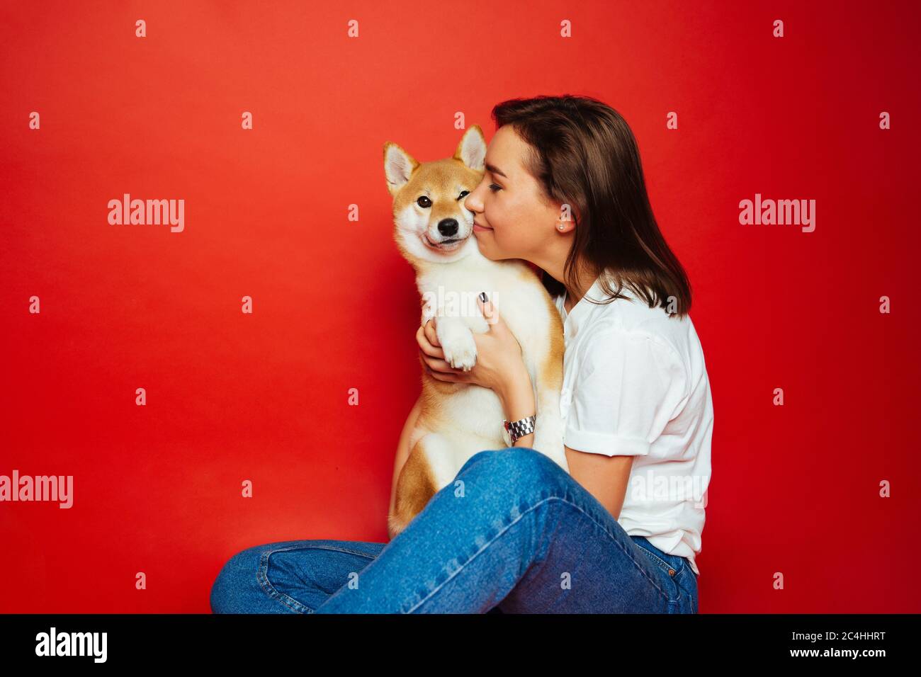 Cute brunette woman in white t shirt and jeans holding and embracing Shiba Inu dog on plane red background. Love to the animals, pets concept Stock Photo