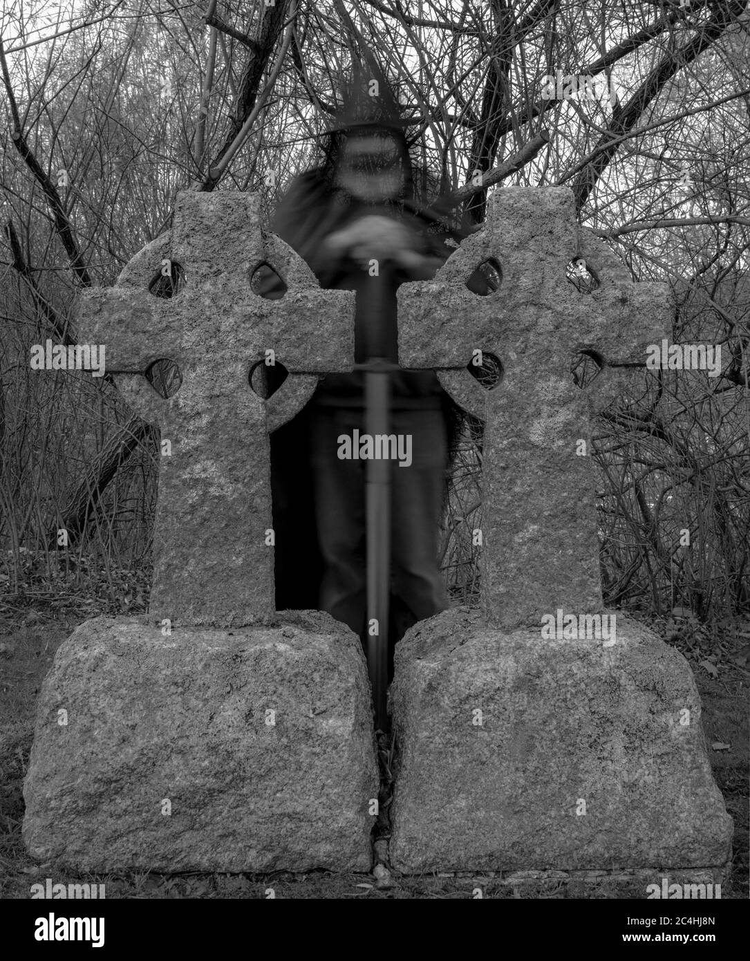 A ghost of a hooded knight with a sword standing behind Celtic grave markers at dusk. Black and white. Stock Photo