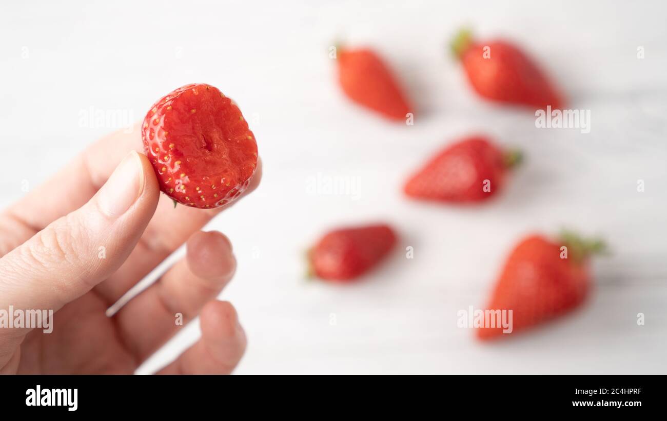 Hand holding bitten strawberry on white wooden background. Summer berry pleasure. Scattered strawberries Stock Photo