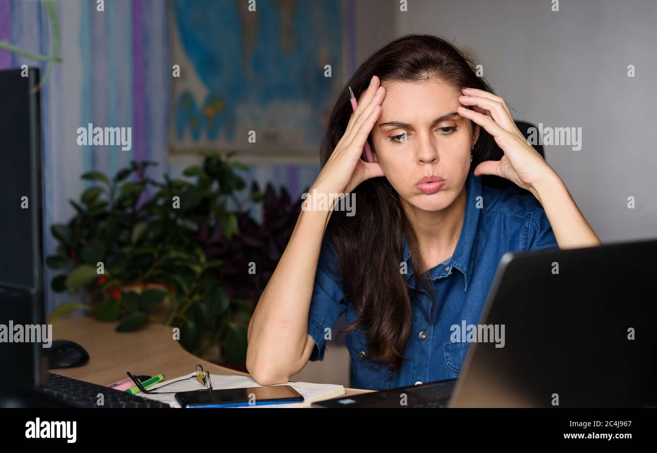 Too much work stressed young woman sitting at her desk in front of computer on world map background. Isolated home because of the corona virus Stock Photo
