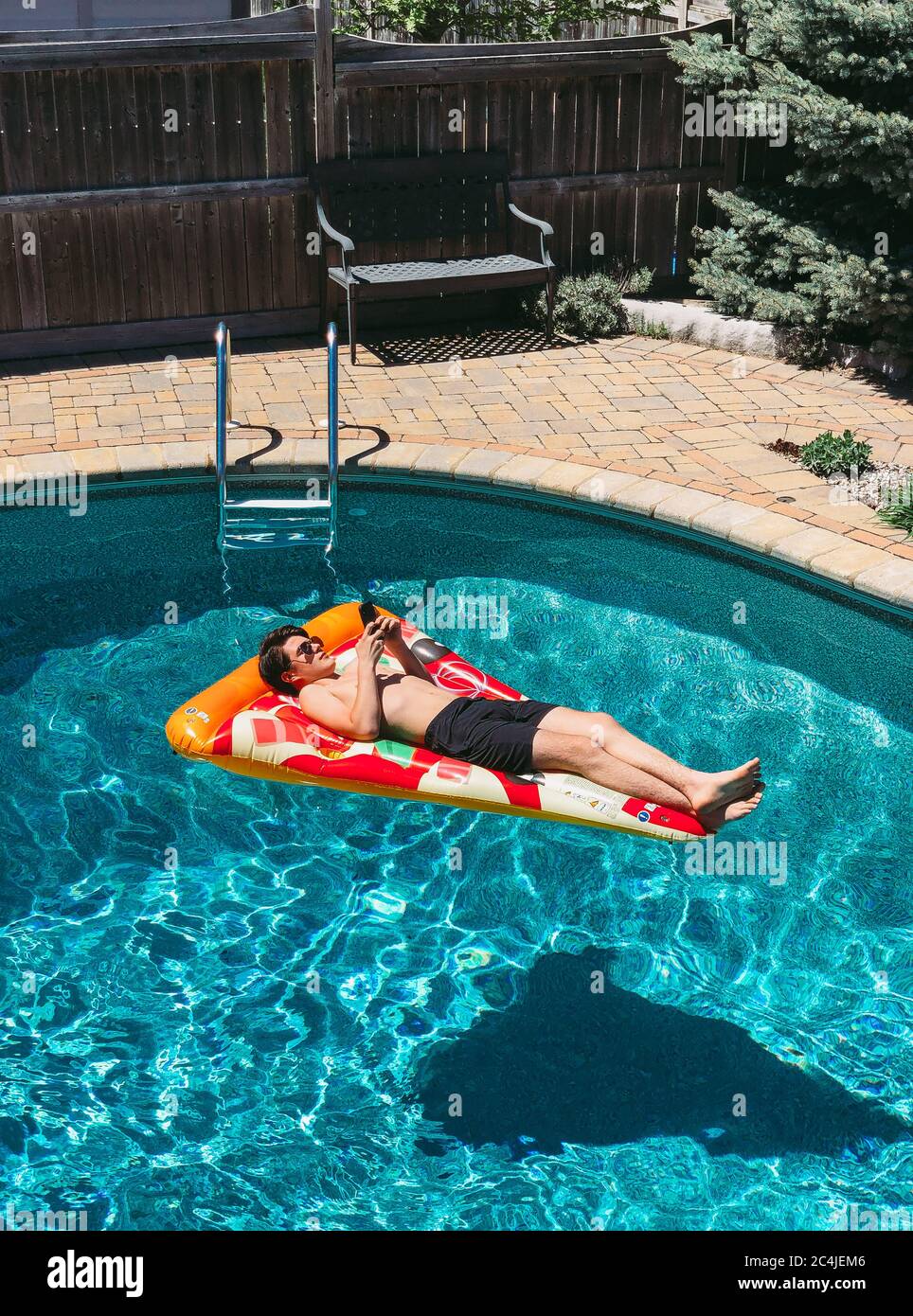 Teenage boy laying on pizza floatie in backyard pool on his cellphone 
