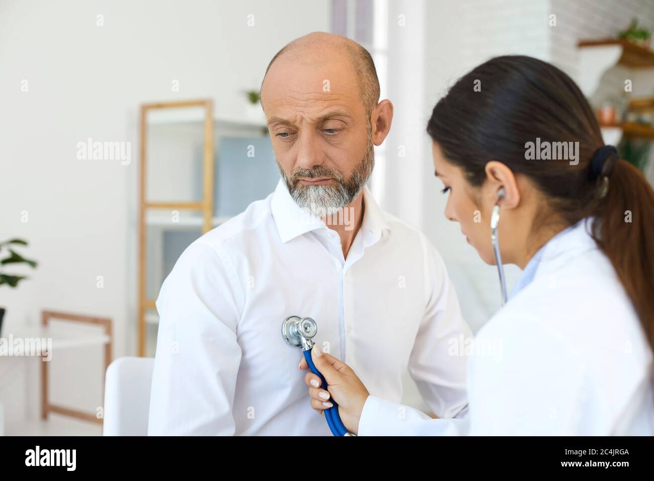 Young female cardiologist listening to senior man's heartbeat at clinic. Doctor examining elderly patient at office Stock Photo