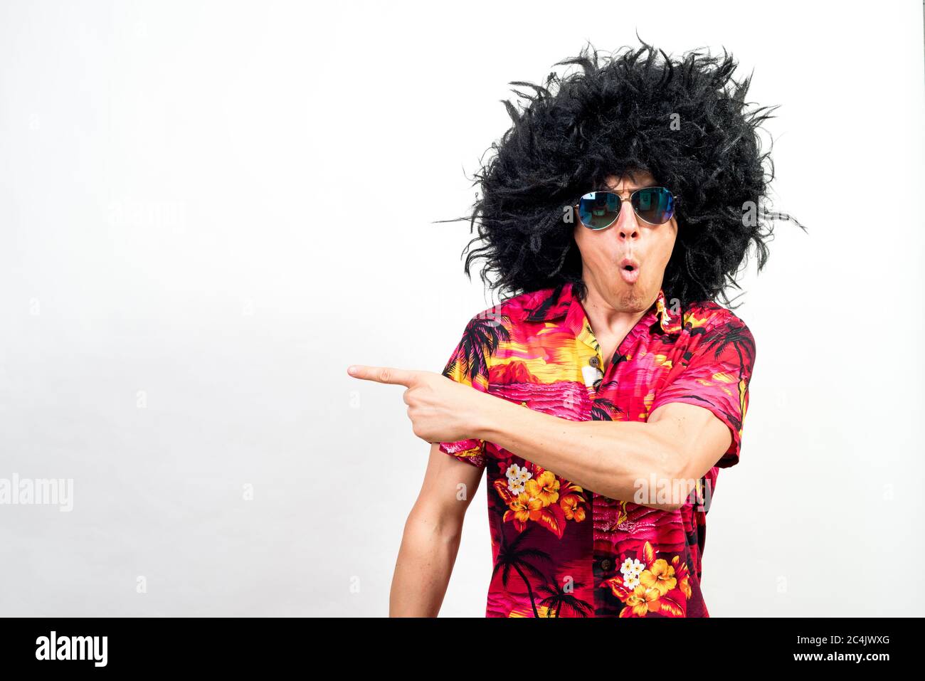 Man in afro wig, sunglasses and hawaiian shirt, pointing aside very surprised. Mid shot. White background. Stock Photo