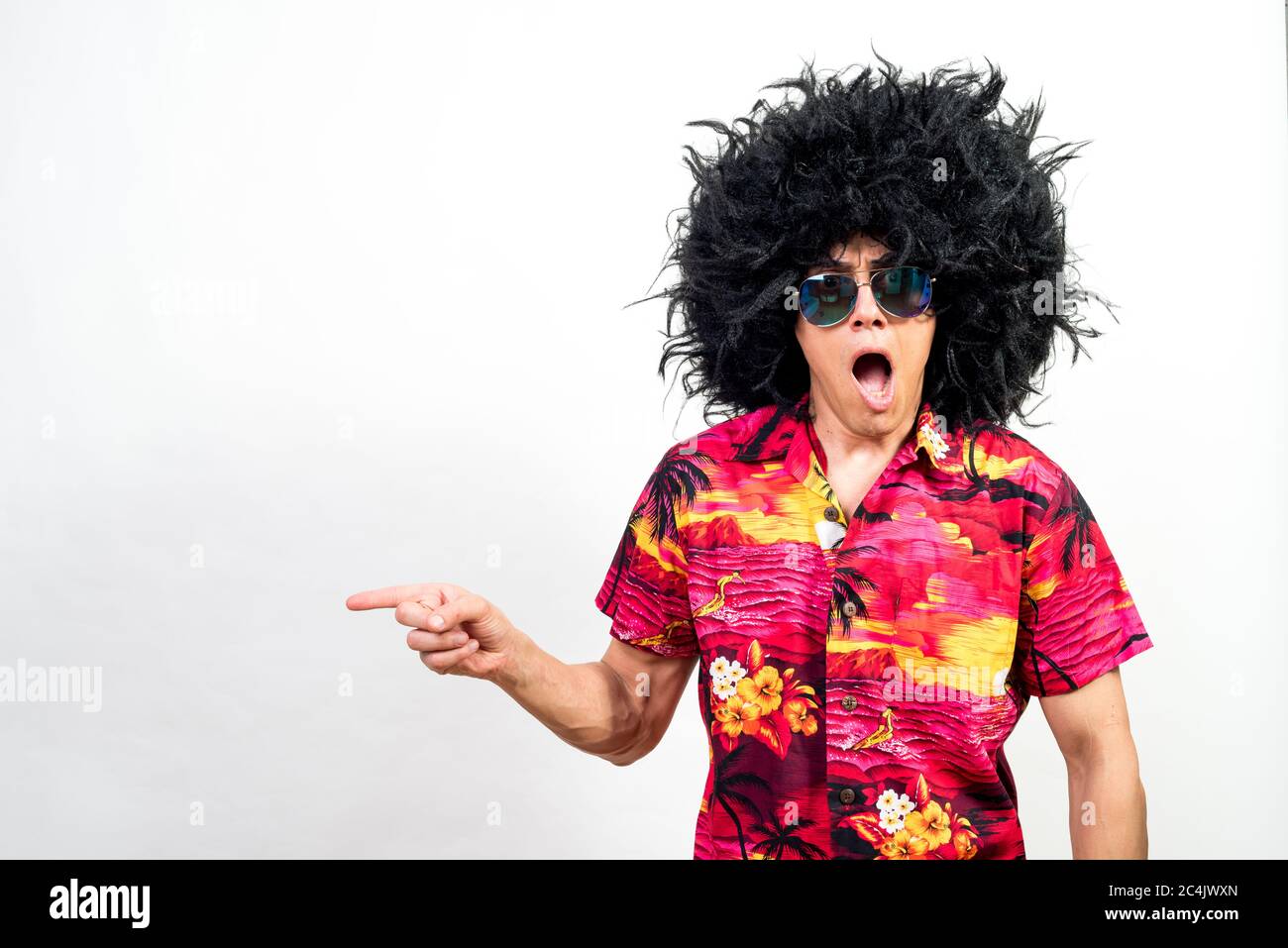 Man in afro wig, sunglasses and hawaiian shirt, pointing aside very surprised. Mid shot. White background. Stock Photo