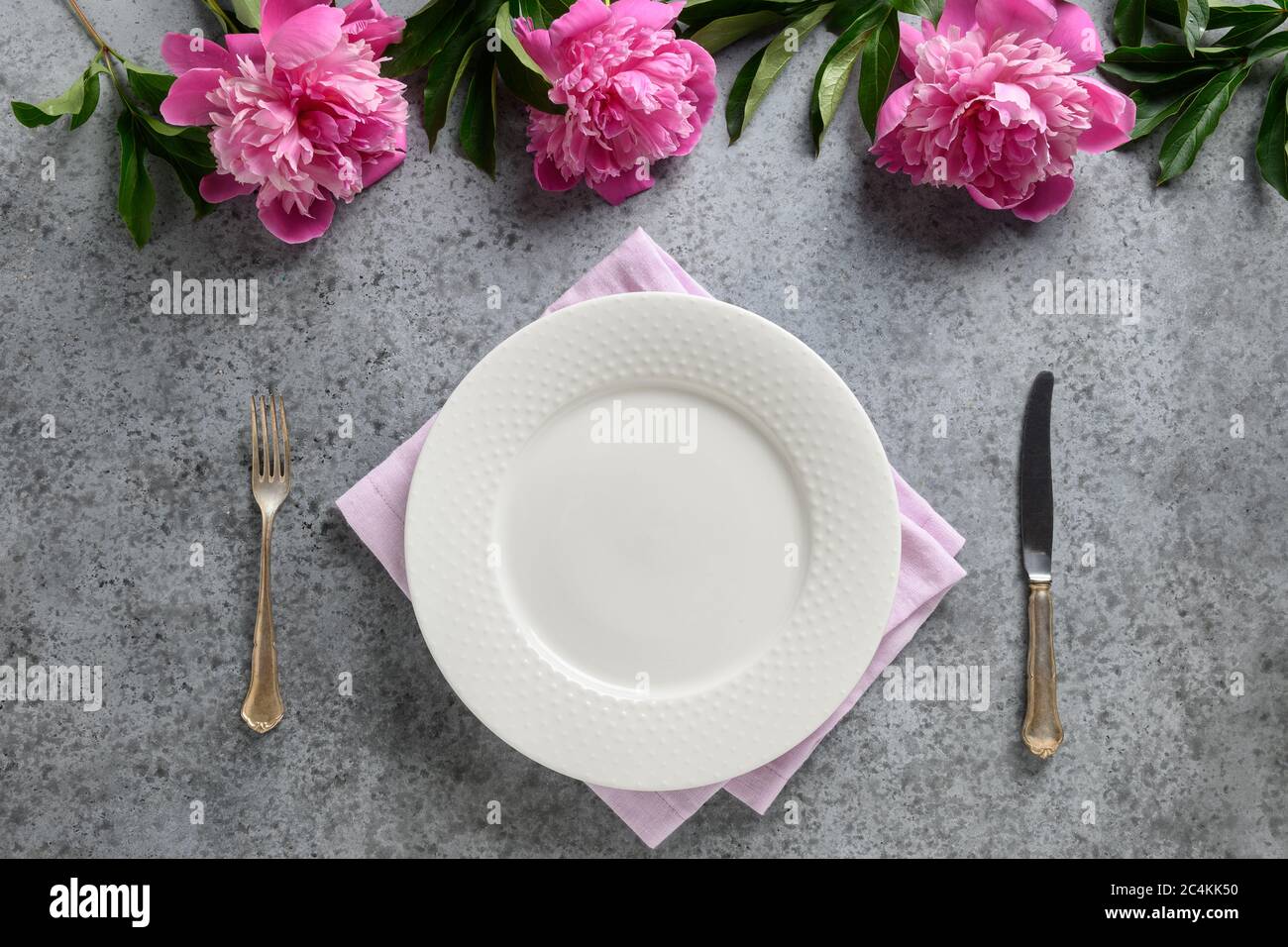 Festive table setting with pink peony, white dishware, silverware on gray. Top view. Stock Photo