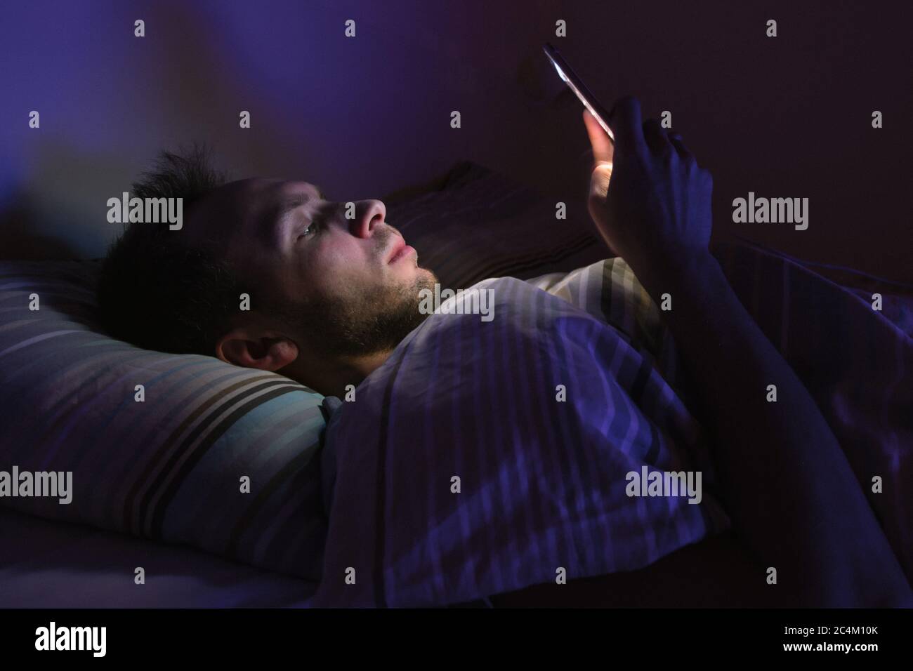Closeup portrait of young sleepy exhausted man lying in bed using smartphone at late night, can not sleep.Insomnia, nomophobia, sleep disorder Stock Photo