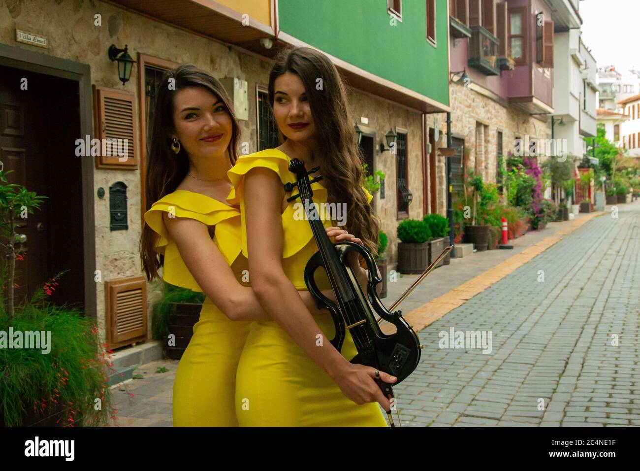 Two beautiful twin sisters violinists in yellow concert dresses are posing with electric violins
