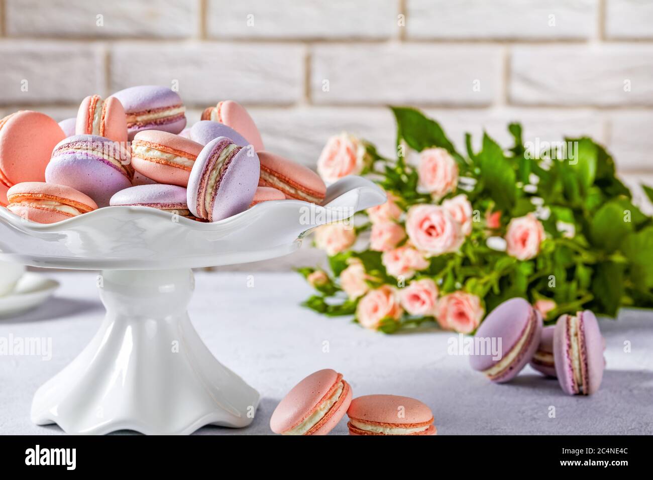macarons on a white porcelain cake stand.  beautiful bouquet of roses and  brick wall at the background, close-up Stock Photo