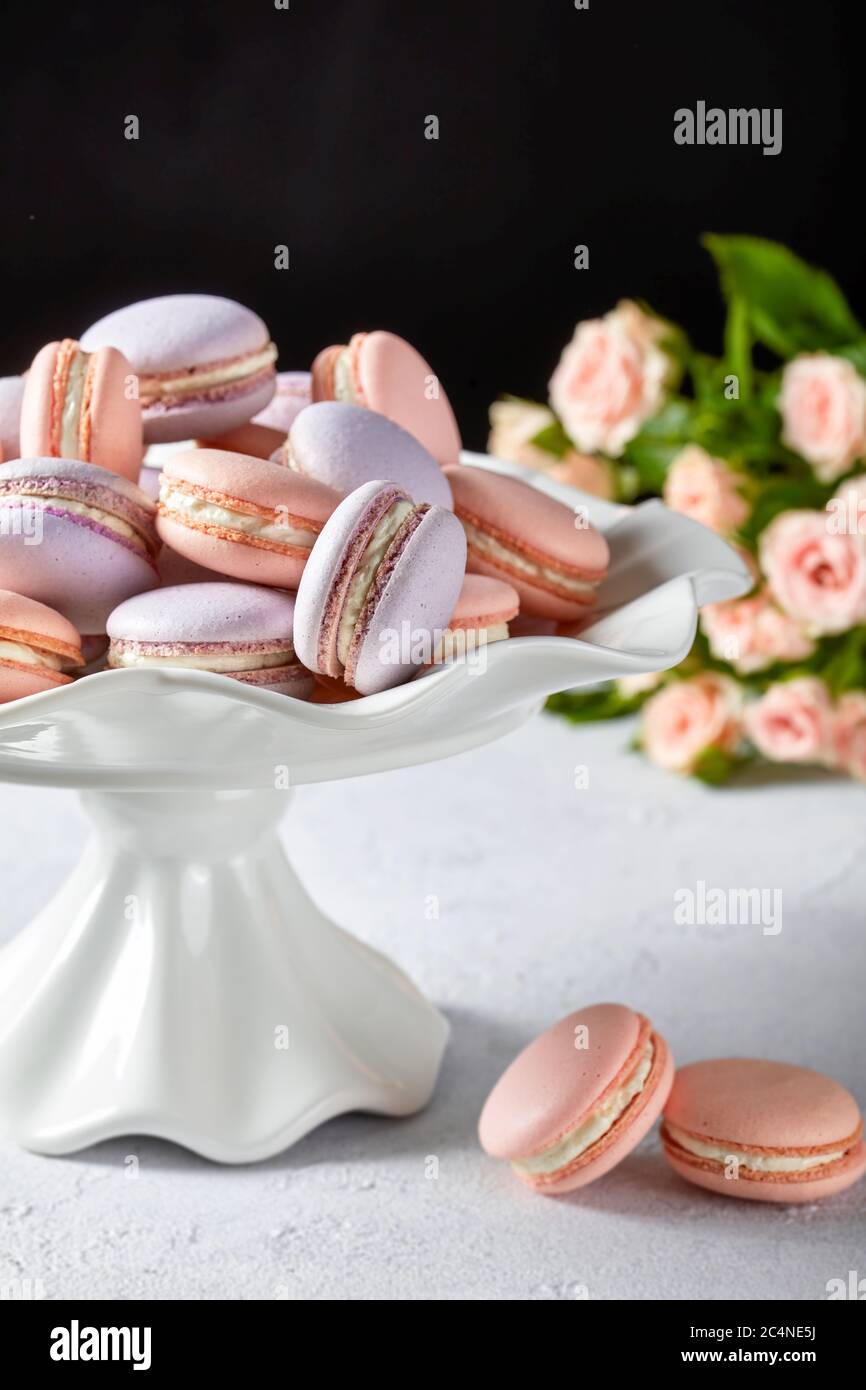 pink and lavender macarons on a white porcelain cake stand with beautiful bouquet of roses at the black background, vertical view, close-up Stock Photo