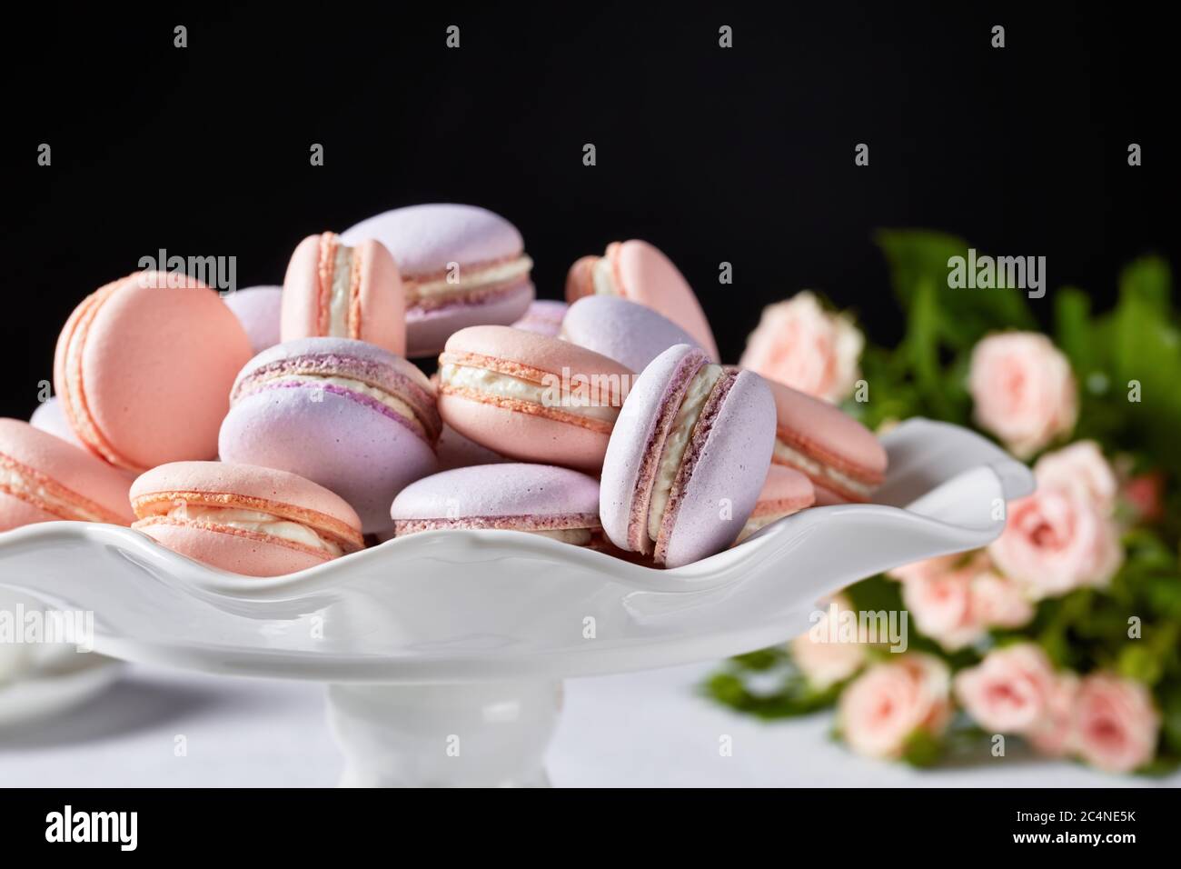 macarons on a white platter with beautiful bouquet of roses at the black background, horizontal view, close-up, free space Stock Photo
