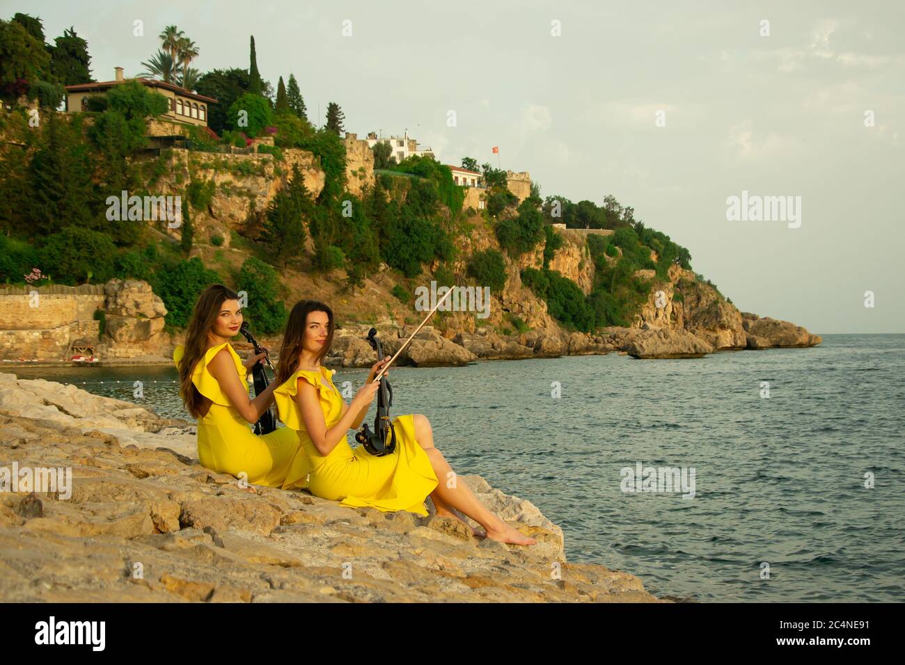 Two beautiful twin sisters violinists in yellow concert dresses are sitting on rocks by the