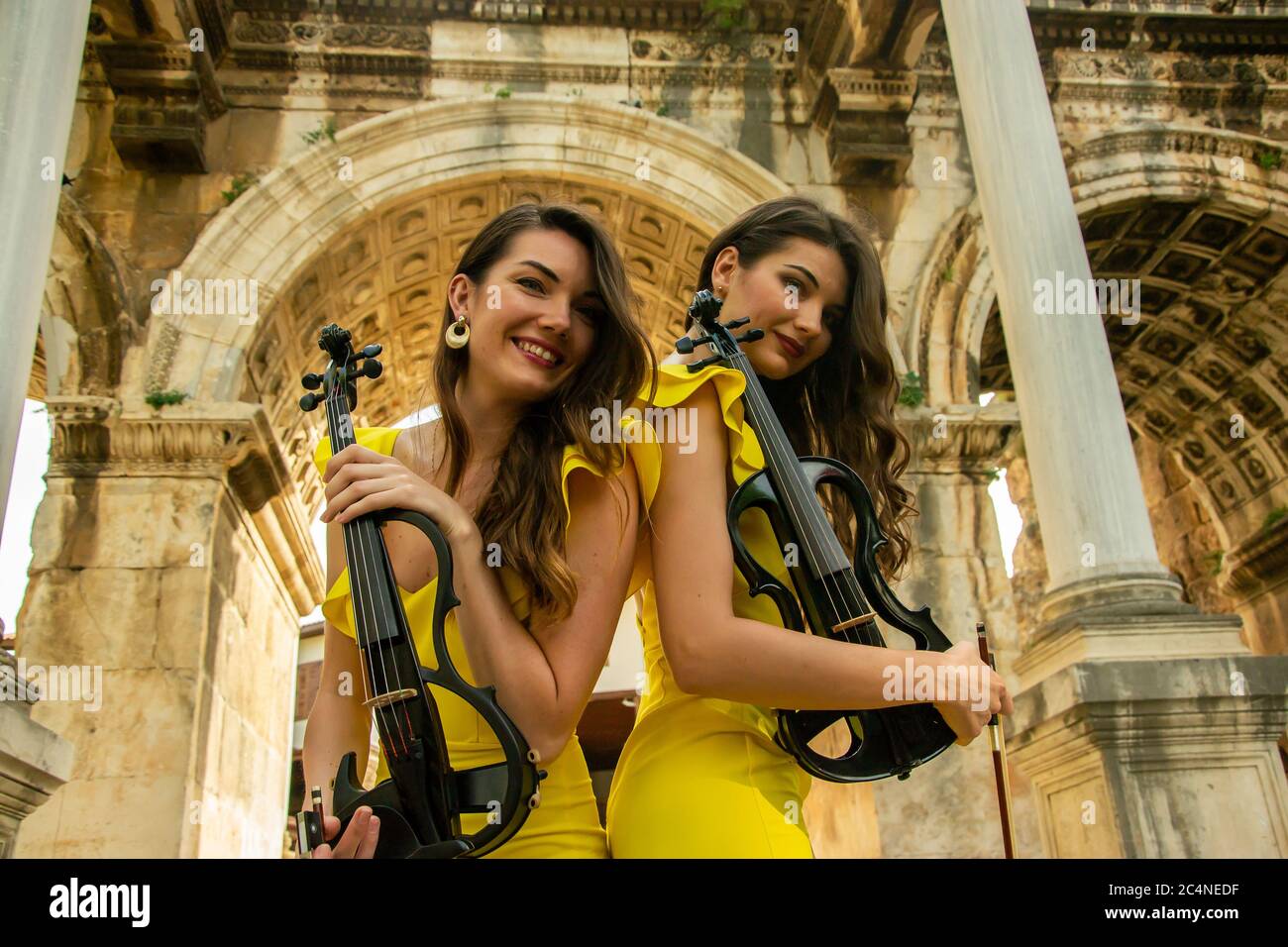 Two beautiful twin sisters violinists in yellow concert dresses are posing with electric violins