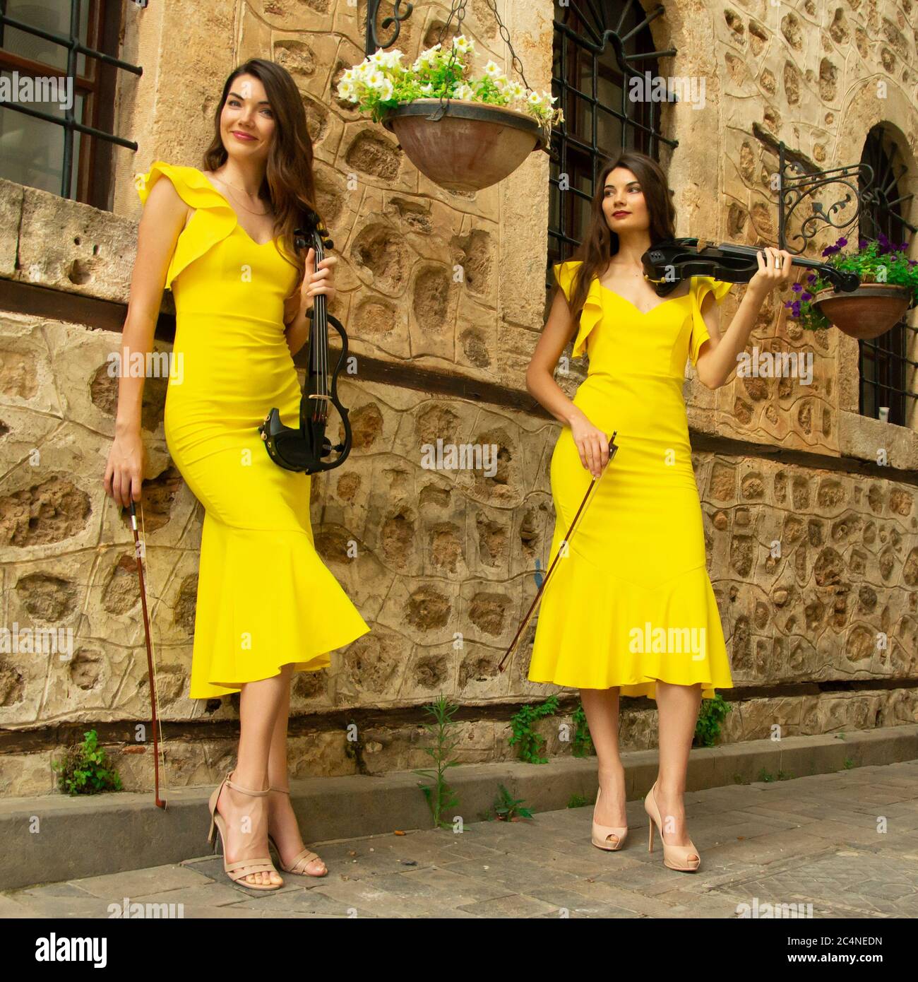 Two beautiful twin sisters violinists in yellow concert dresses are posing with electric violins