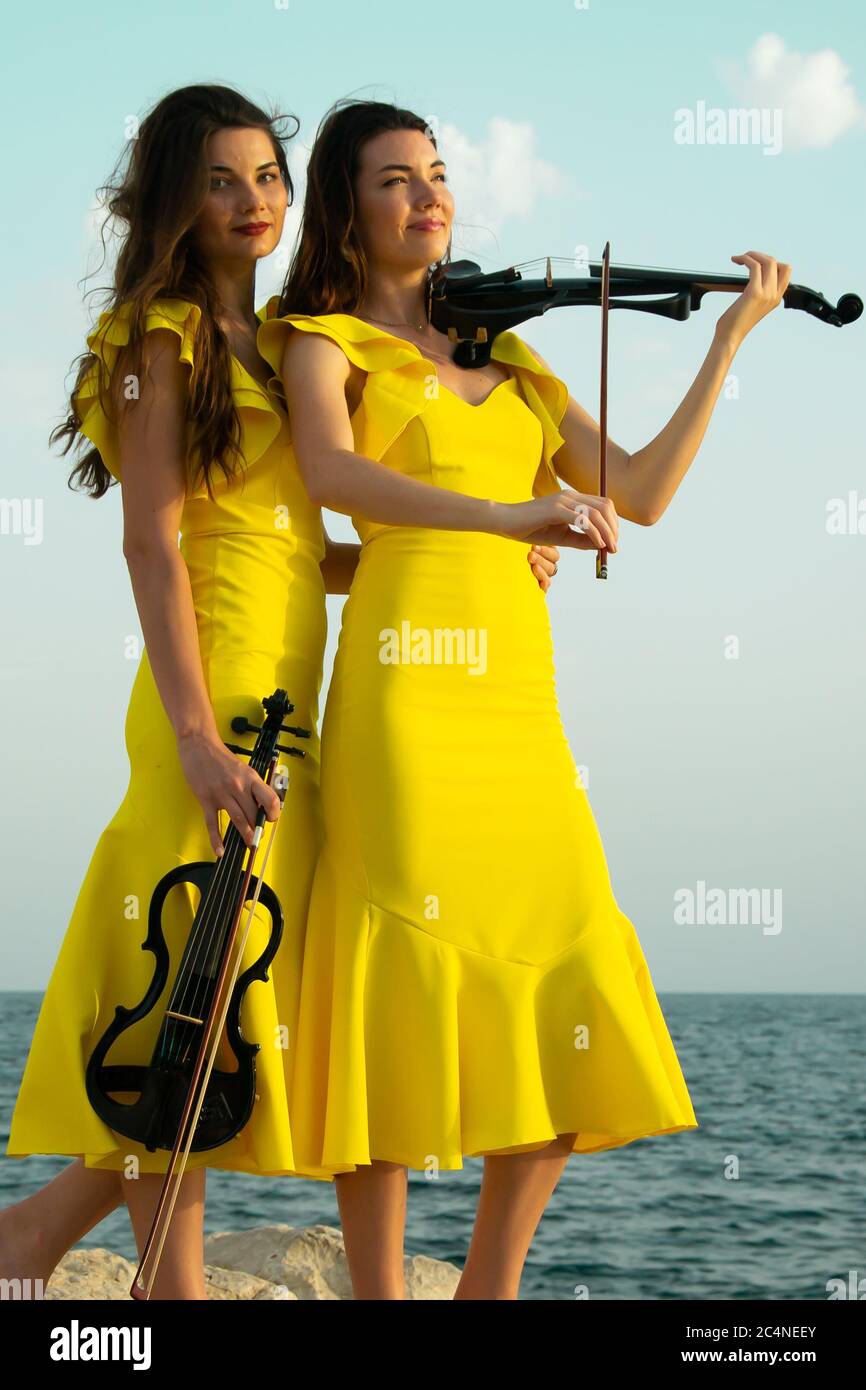 Two beautiful girls violinists in yellow concert dresses are playing electric violins with