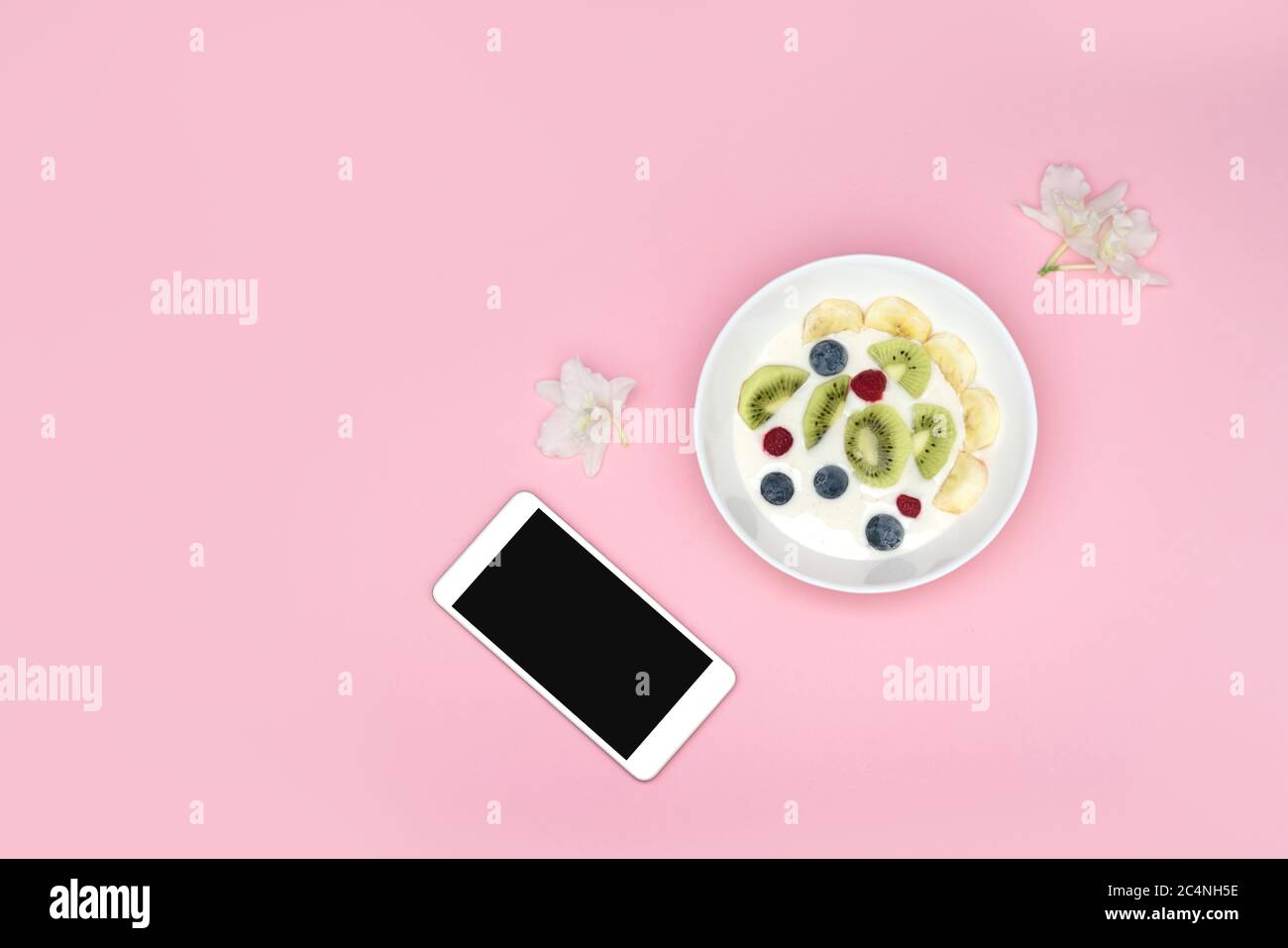 A smoothie bowl with superfoods and a phone on a pink background. Yogurt with blueberries, kiwi, banana and cereal. View from above. Stock Photo
