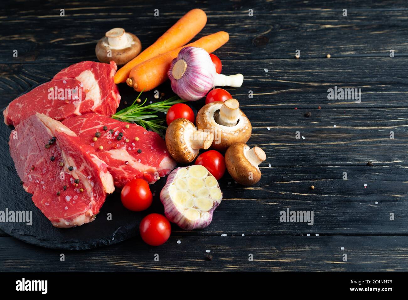 Three pieces of juicy raw beef with rosemary on a stone cutting board on a black wooden table background. Meat for barbecue or grill with vegetables pepper and salt seasoning Stock Photo