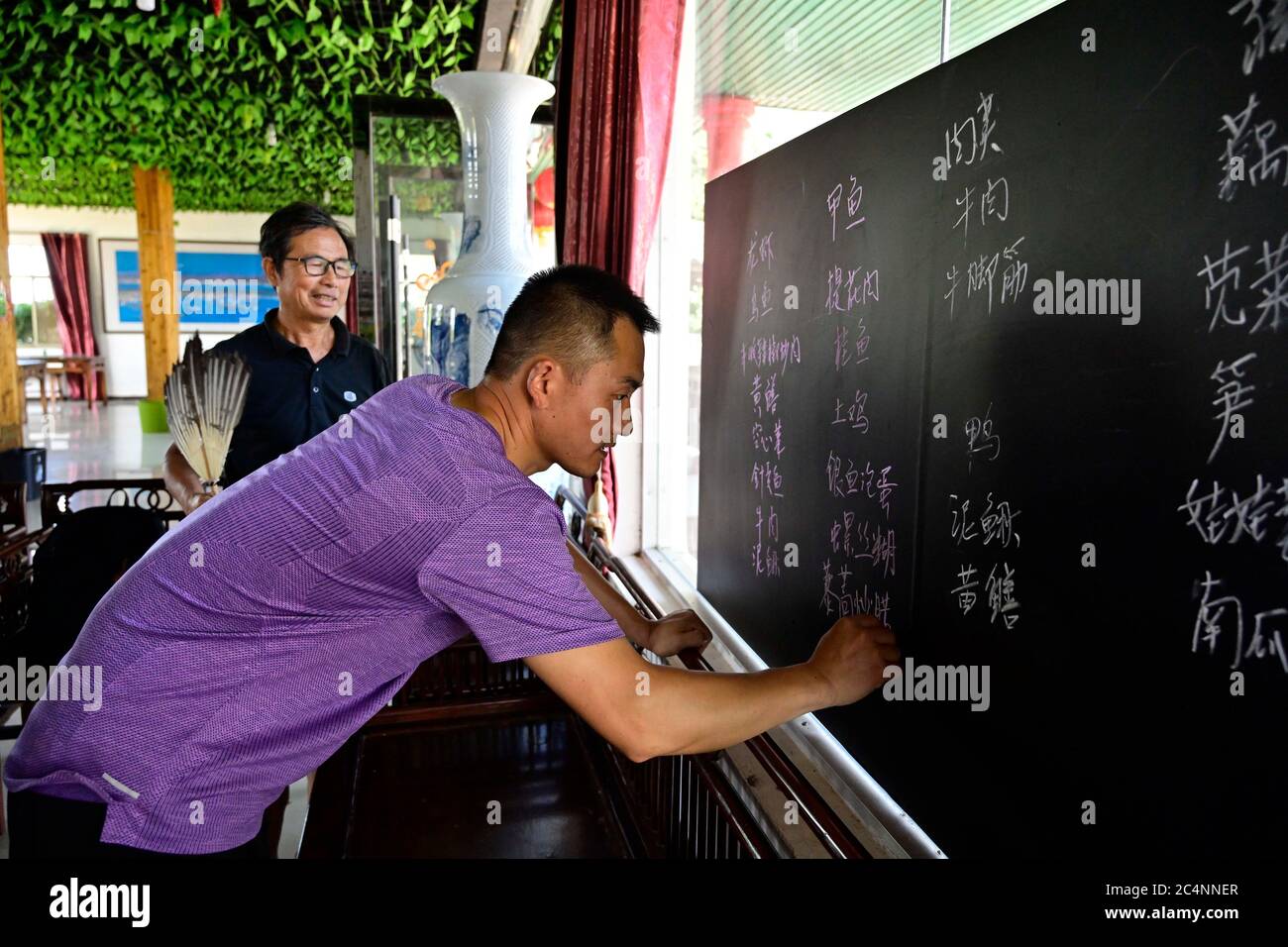 (200628) -- NANCHANG, June 28, 2020 (Xinhua) -- Zhu Yicai (Rear) and his son make a daily menu for guests at his farmhouse in the Poyang Lake scenic area in Yugan County, east China's Jiangxi Province, June 16, 2020. Zhu Yicai gave up his fishing business in 2012 due to the worsening aquatic environment in the Poyang Lake. In 2018, Zhu was running a family farm when he noticed that lots of tourists came to the lake to watch migratory birds. Zhu then captured the business opportunity and opened a 1,800-square-meter farmhouse, which can accommodate more than 200 people dining at the same time. C Stock Photo