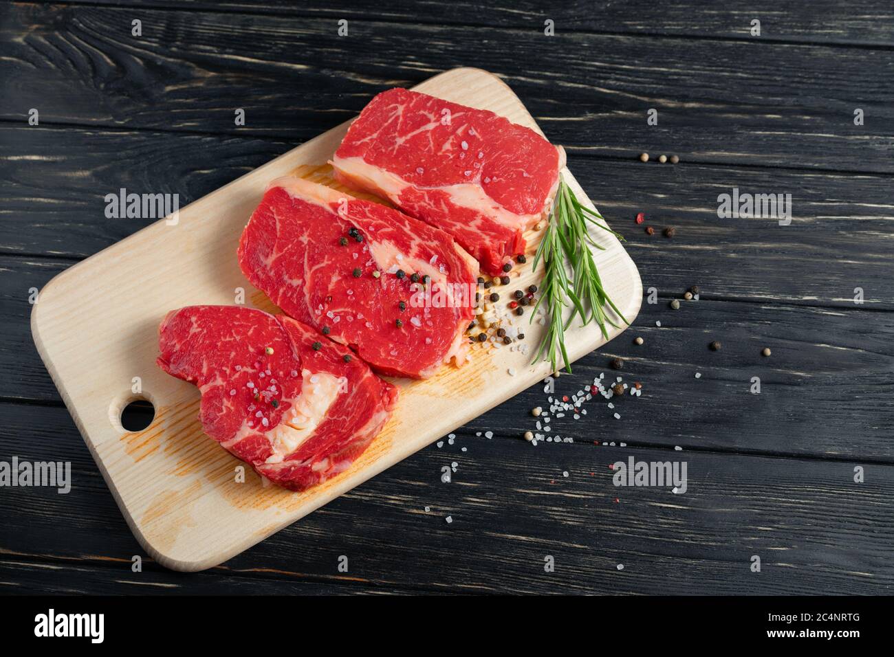 Three pieces of juicy raw beef with rosemary on a cutting board on a black wooden table background. Meat for barbecue or grill sprinkled with pepper and salt seasoning Stock Photo