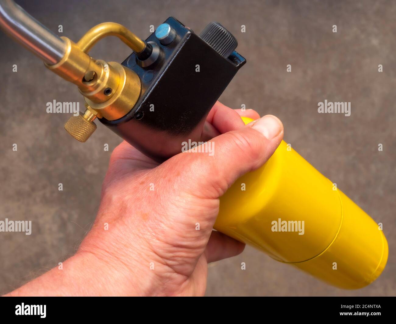 Closeup POV shot of a man’s hand holding a traditional blowtorch / blow torch, with a bright yellow gas cylinder / canister. Stock Photo